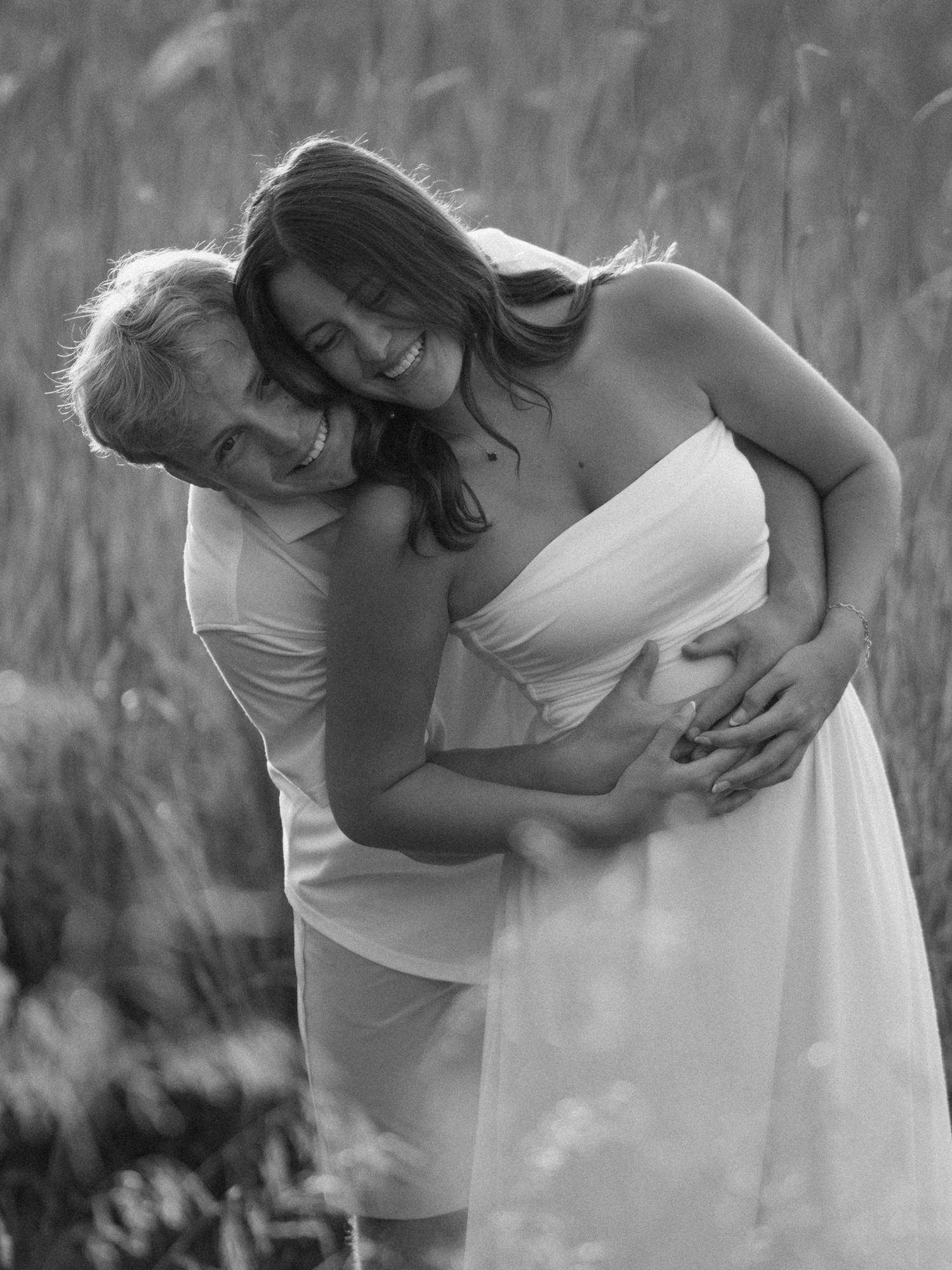 Black and white photo of a couple embracing outdoors, pregnant woman in white dress smiling joyfully.