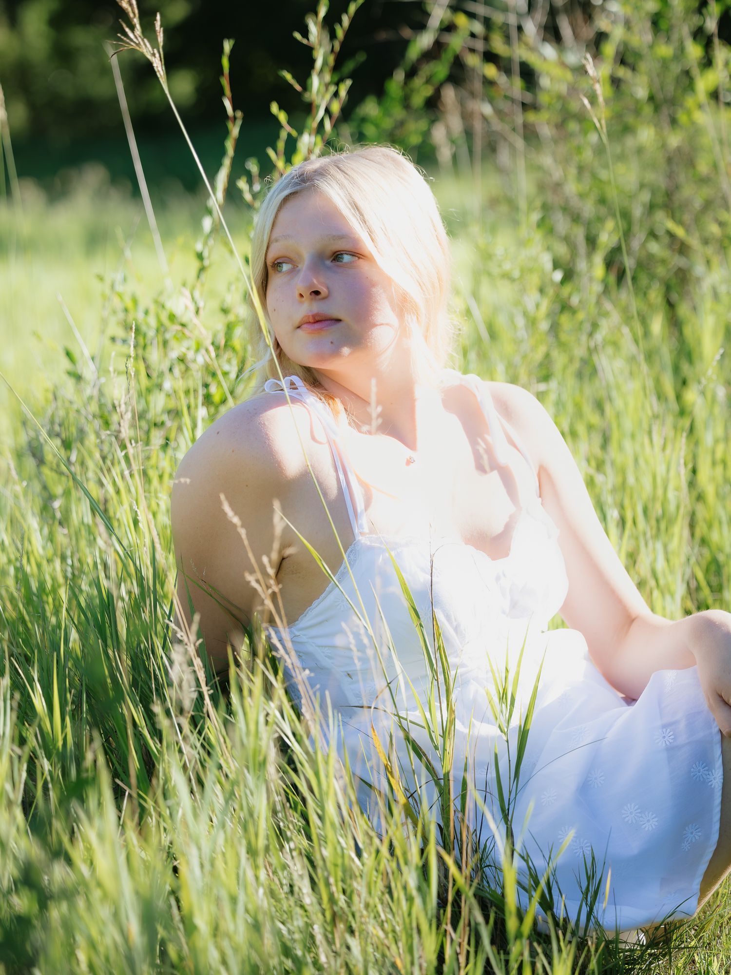 A young blonde woman in a white dress sits peacefully in a sunlit green meadow.