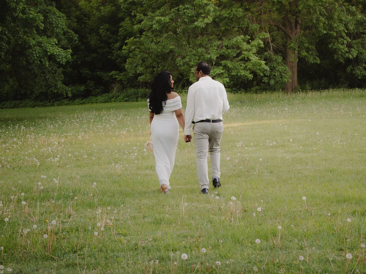 Couple in white outfits walking hand in hand through a lush green meadow.