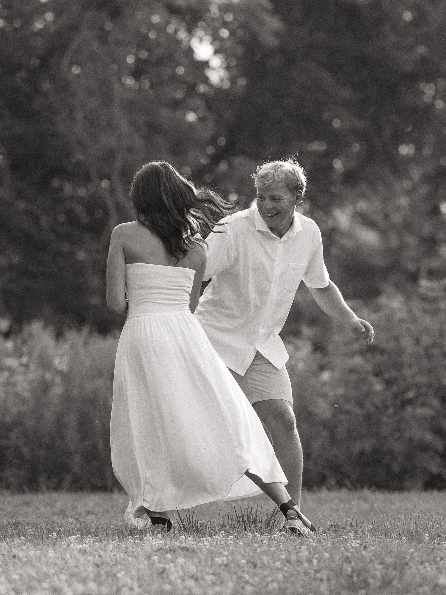 Black and white photo of a joyful couple in white outfits dancing playfully on a grassy lawn with trees in background.