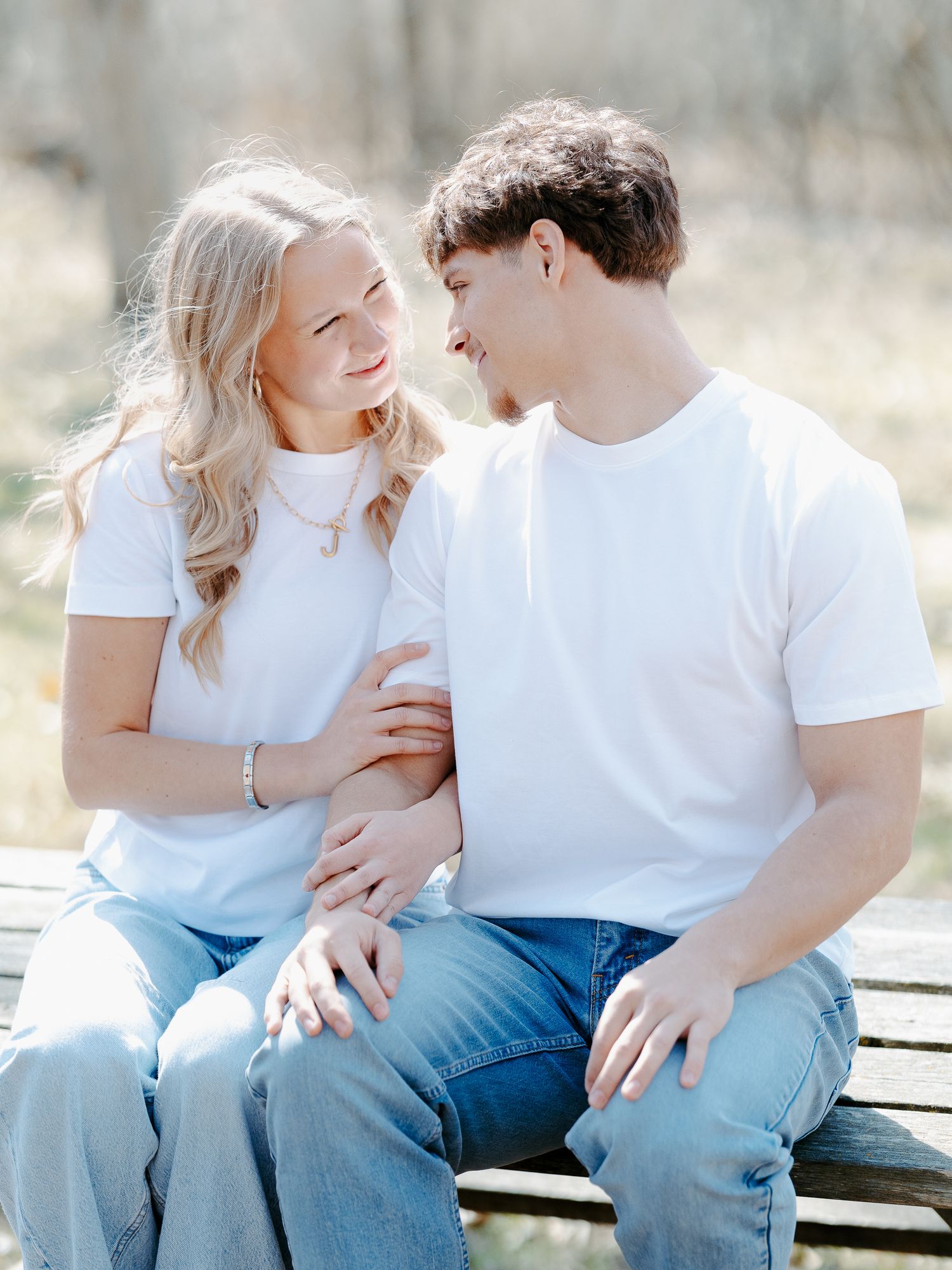 A young couple in matching white tees and jeans sit on a bench, smiling at each other in a sunny outdoor setting.