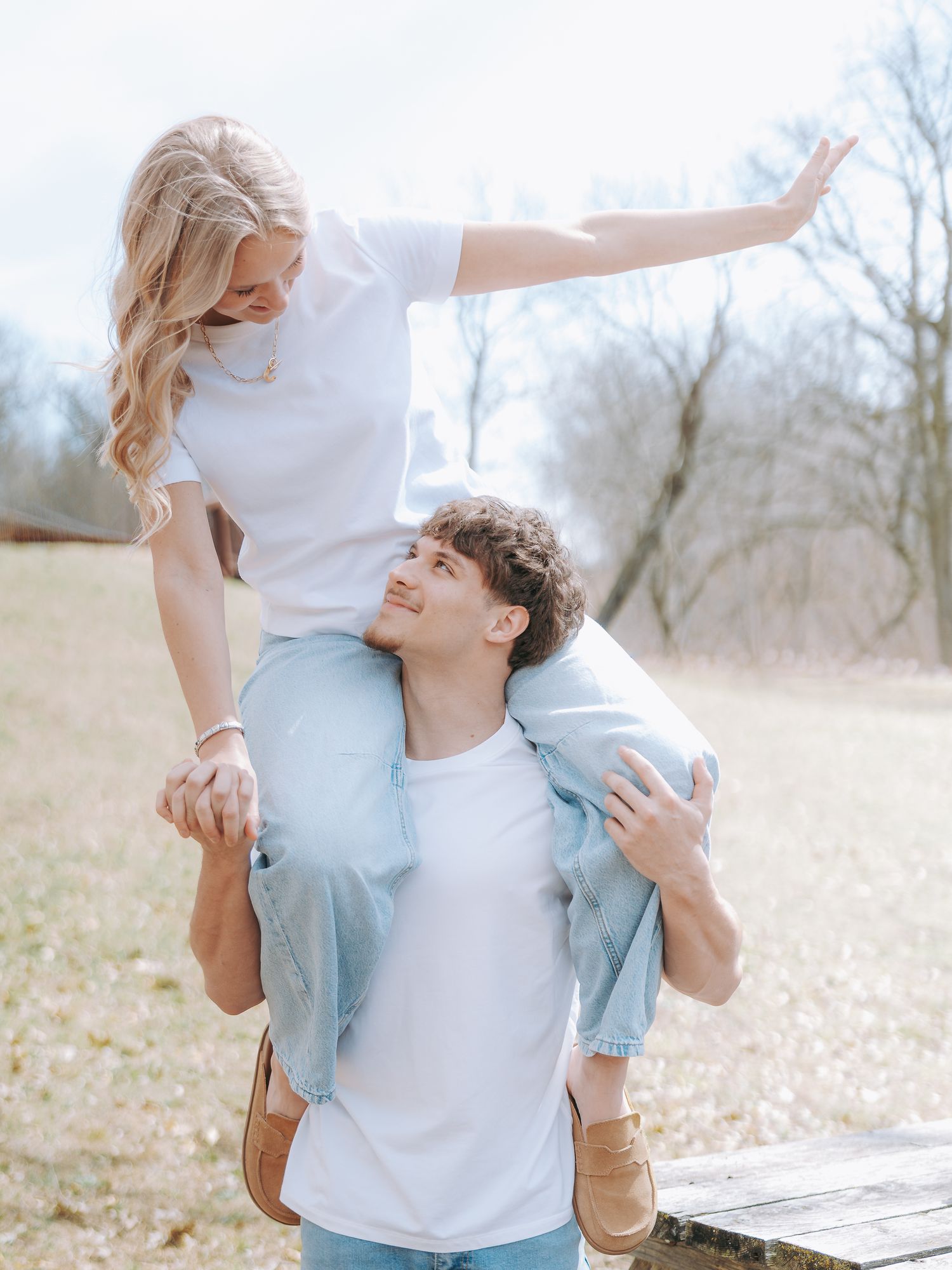 Happy couple in white shirts outdoors, man lifting woman on his shoulders in sunny park setting.