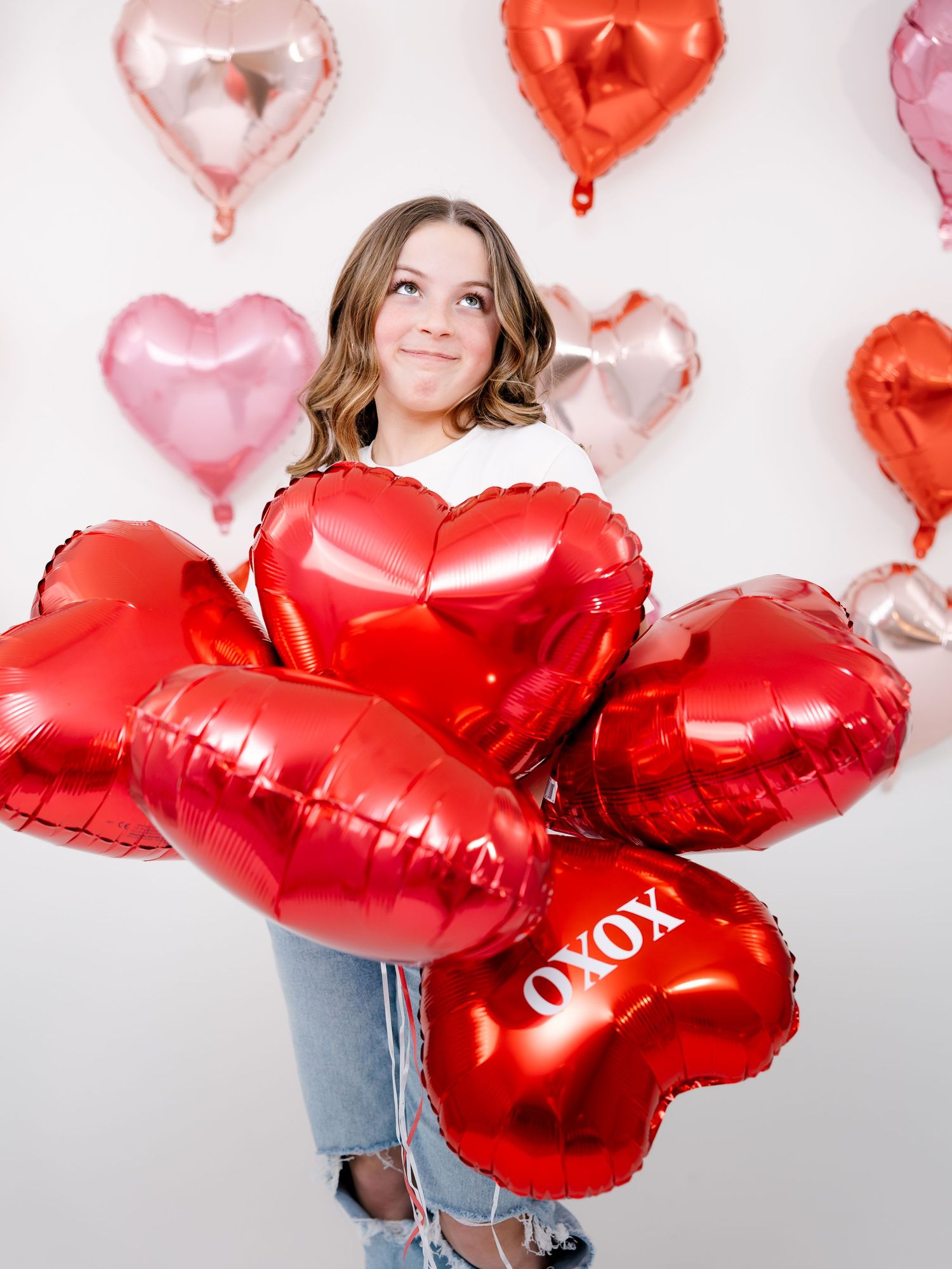 Young girl holding bundle of red XOXO heart balloons with pink and red heart balloons on white wall behind her.