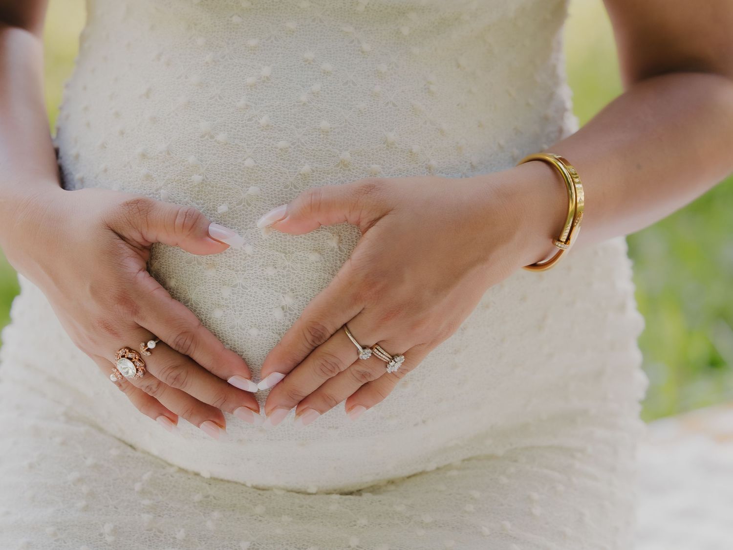A pregnant woman in a white dotted dress forms a heart shape with her hands over her baby bump.