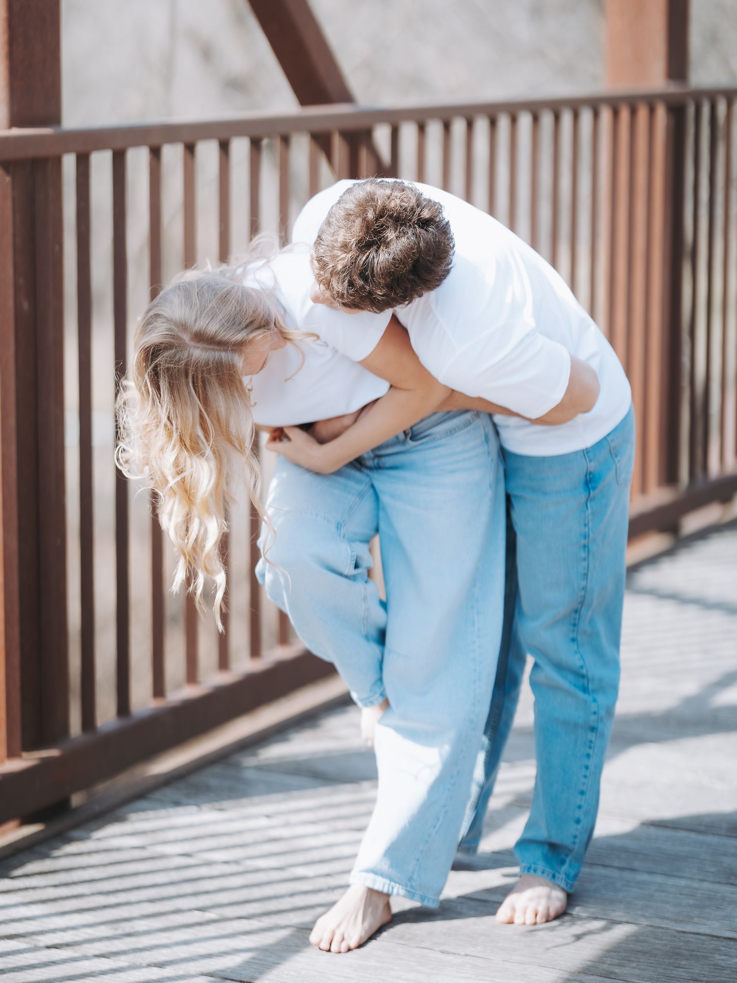 A couple in white shirts and jeans sharing a playful hug on a wooden bridge in sunlight.