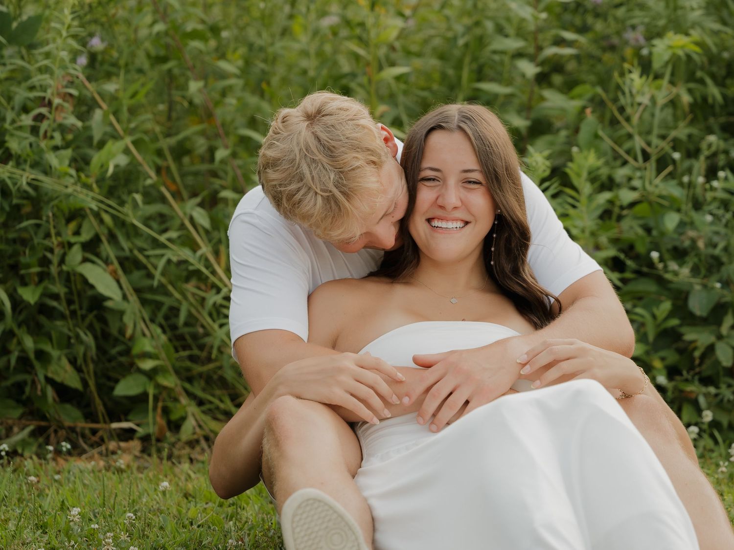 A smiling couple sits together in a lush green field, the man embracing the woman in a white dress.