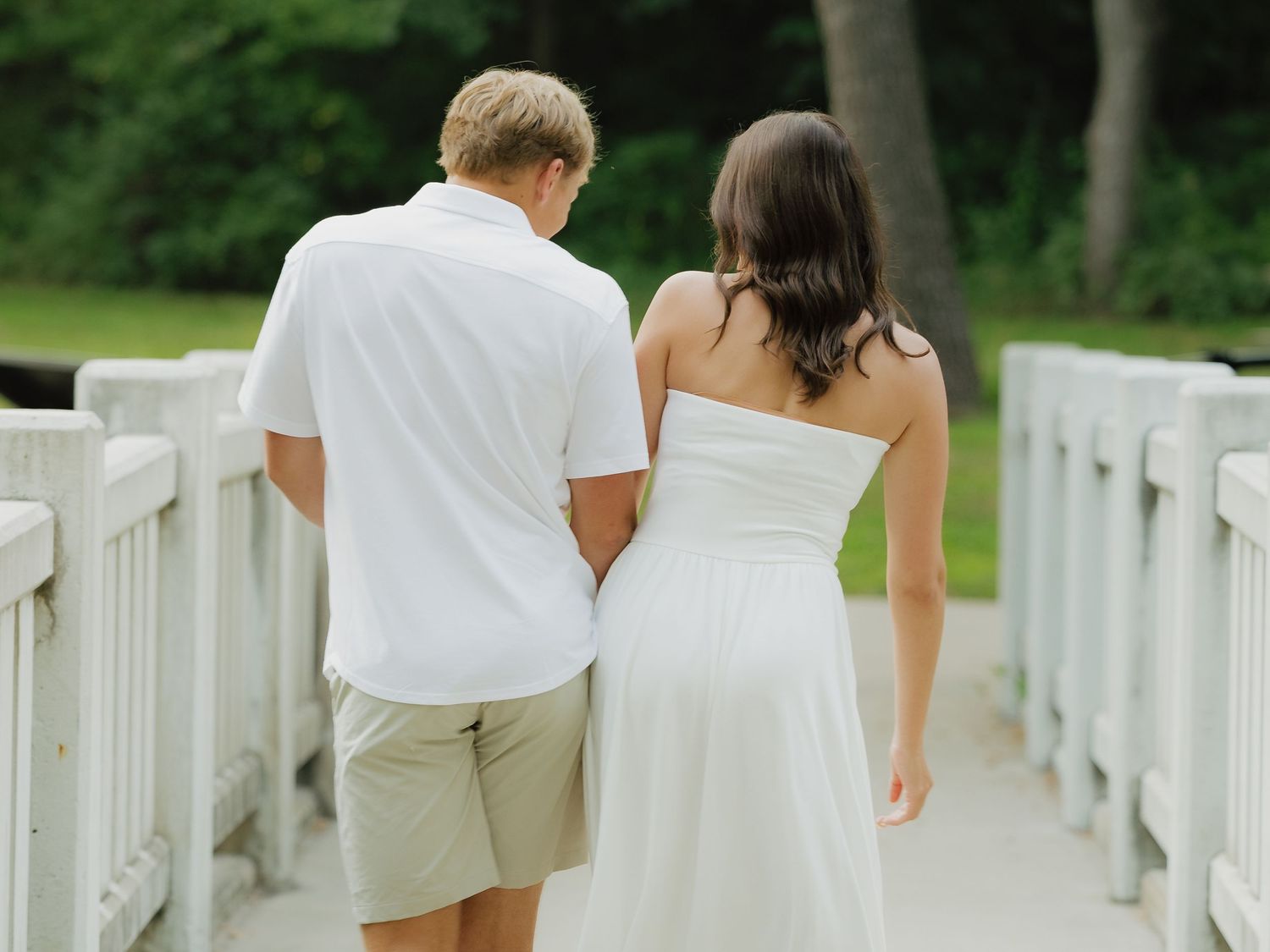 Couple in white outfits walking hand in hand on a bridge surrounded by lush green trees.