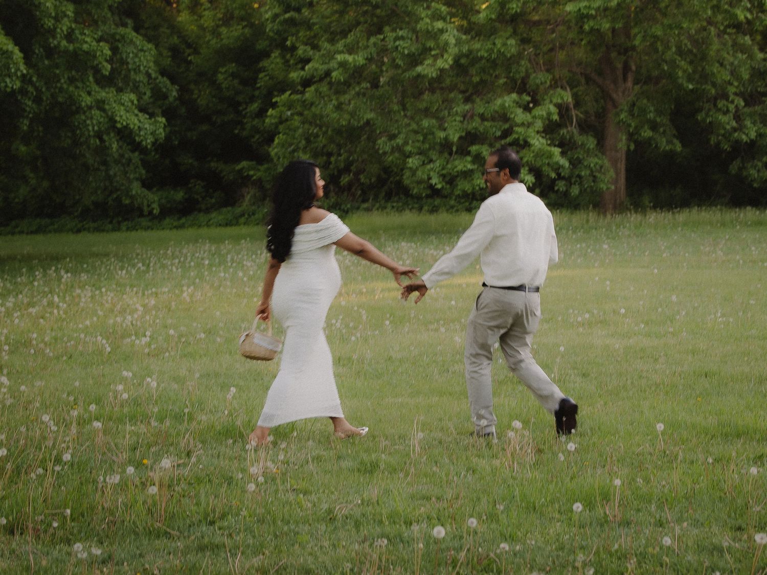 Couple in white outfits holding hands walking through sunny green meadow with wildflowers.