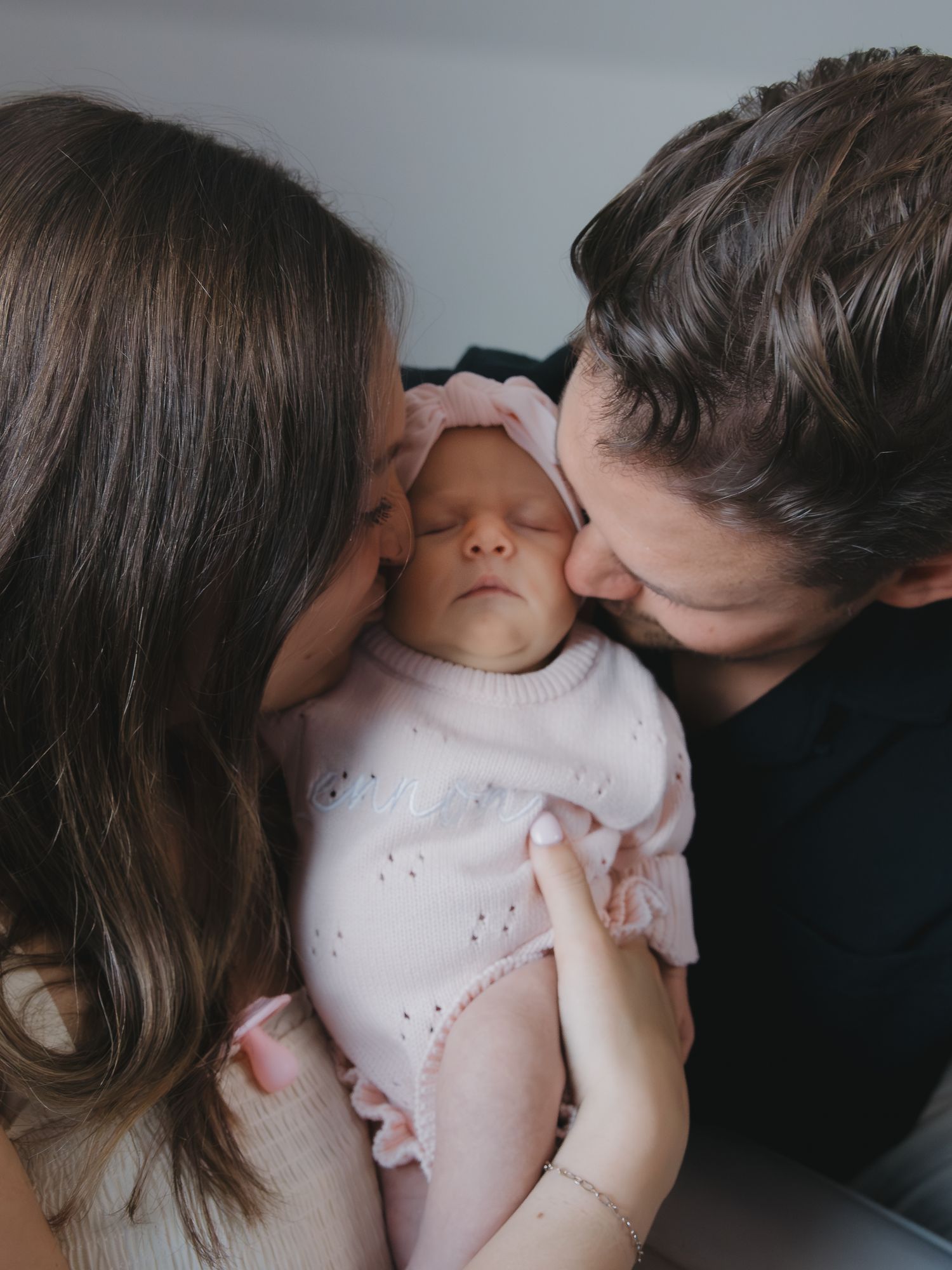Parents tenderly kissing newborn baby in pink outfit on forehead in soft indoor light.