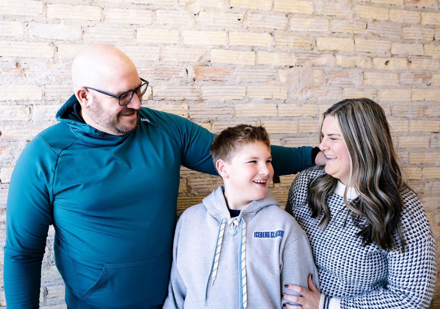 A bald father and brunette mother smile proudly beside their teenage son against a brick wall.