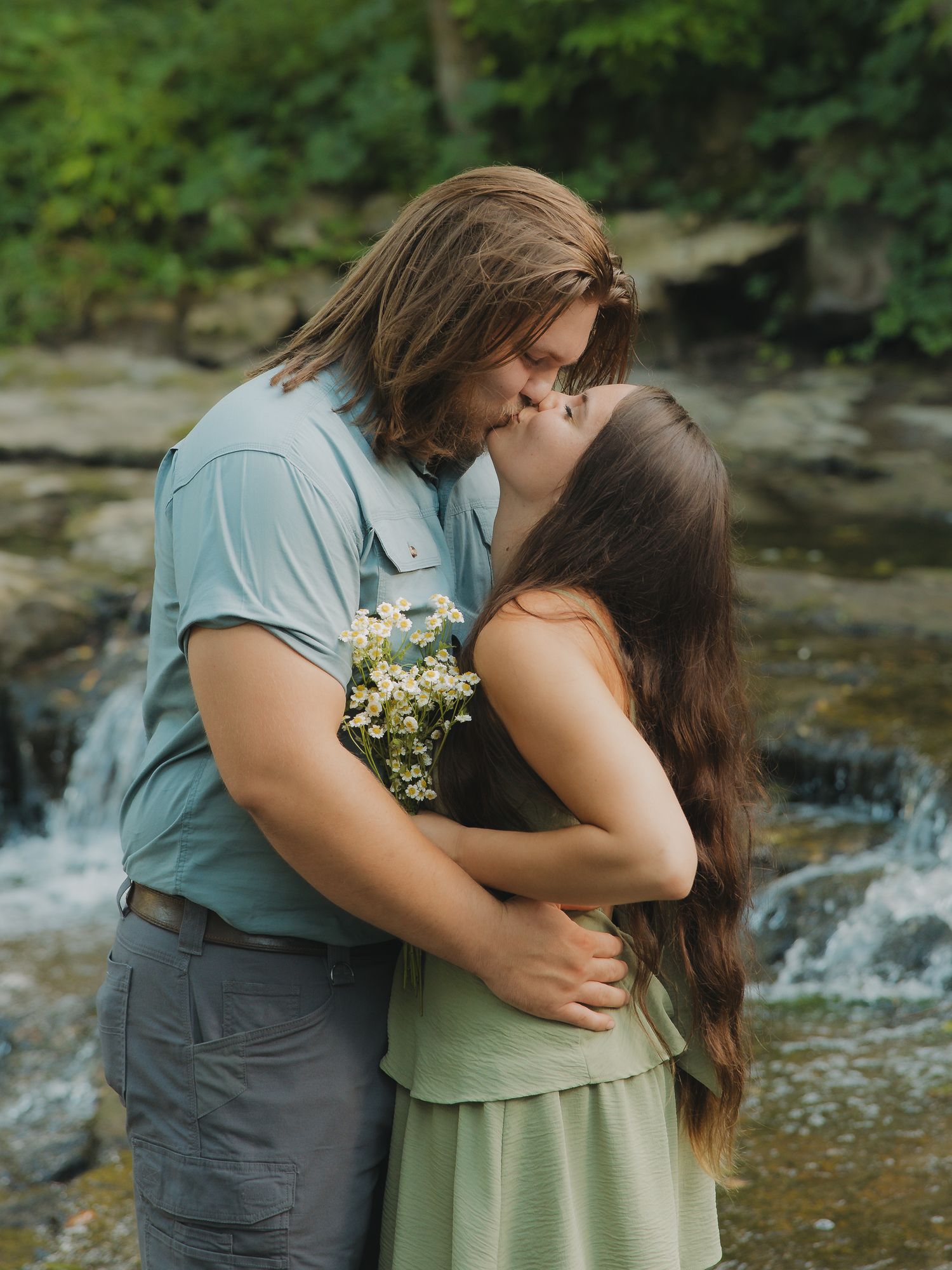 Romantic couple kissing near a waterfall while man holds a bouquet of white wildflowers.