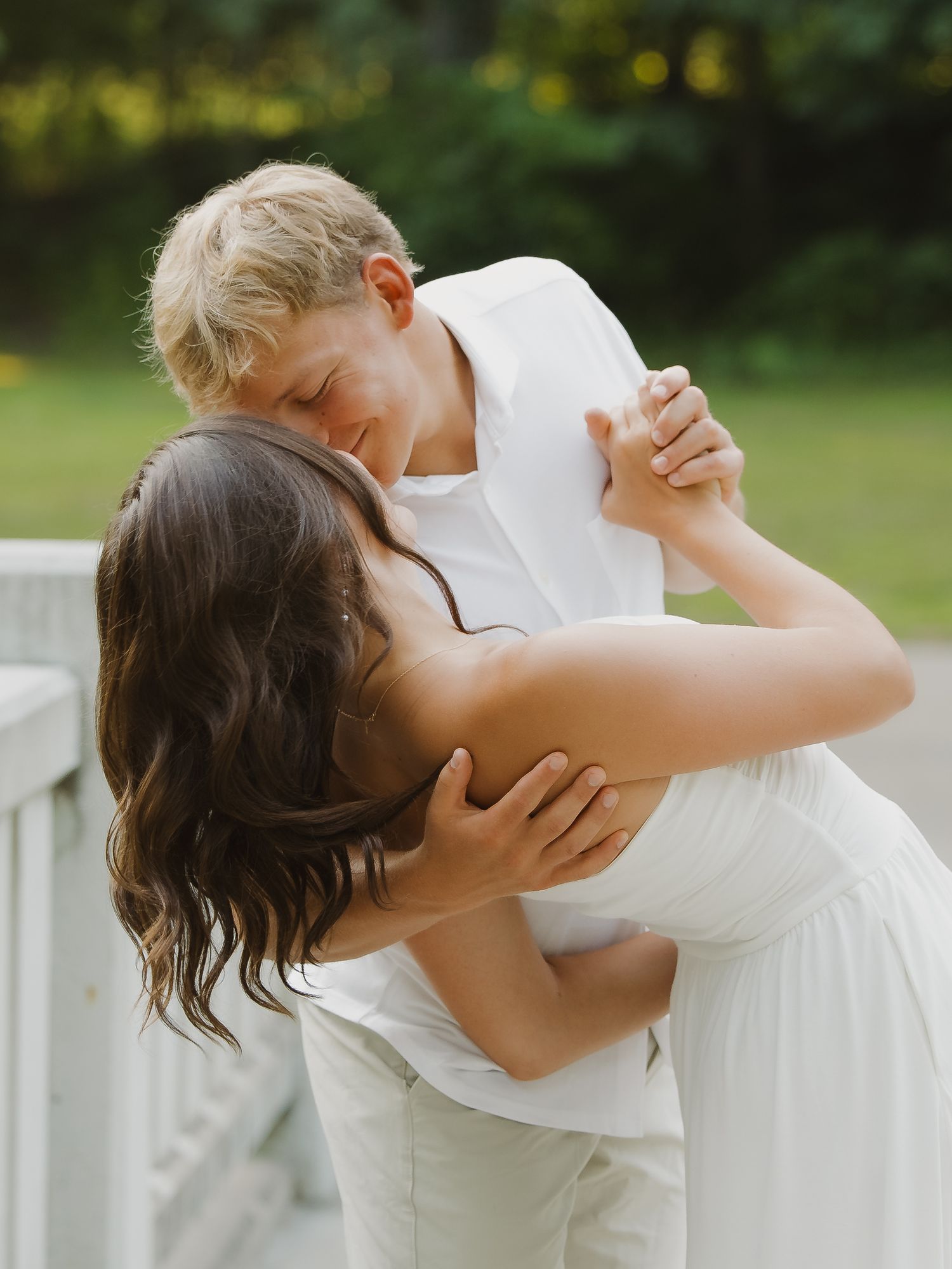 A mother and young blonde boy share a tender embrace while dancing together outdoors in matching white outfits.