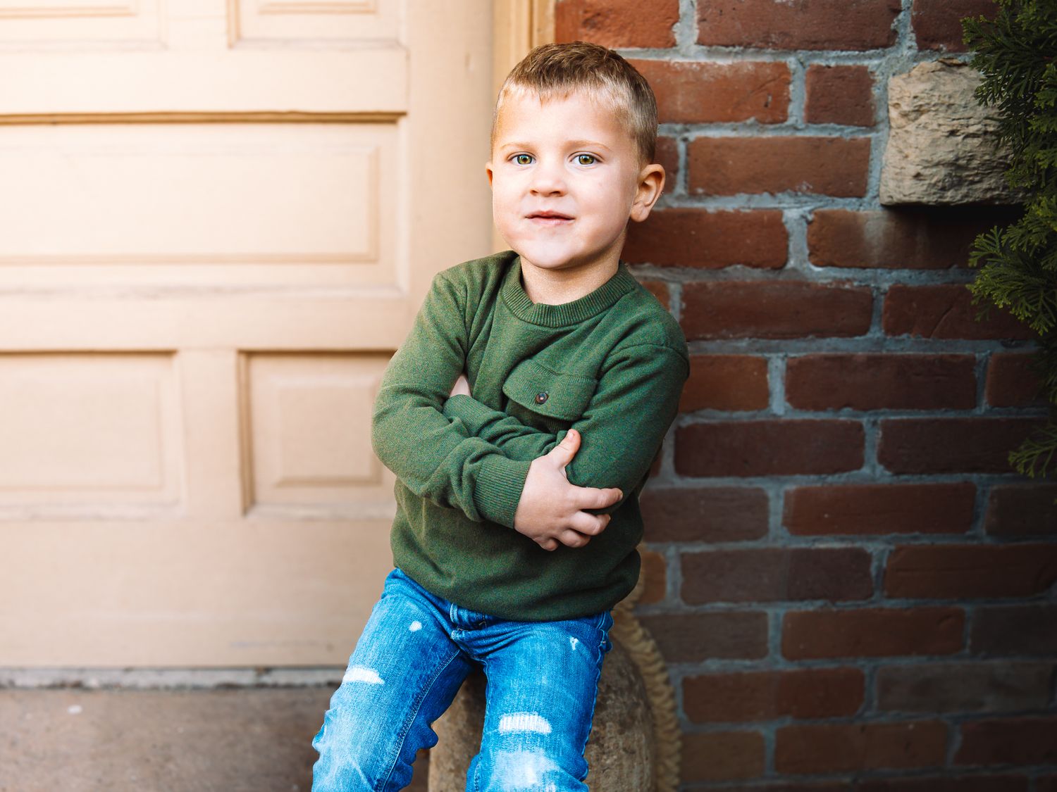 Cute young boy in green shirt and ripped jeans posing confidently by a brick wall.