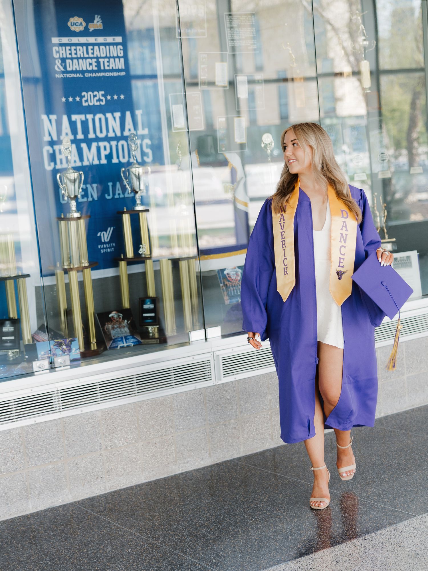 Graduate in purple robe and gold sash walks past 2025 National Champions cheerleading display.