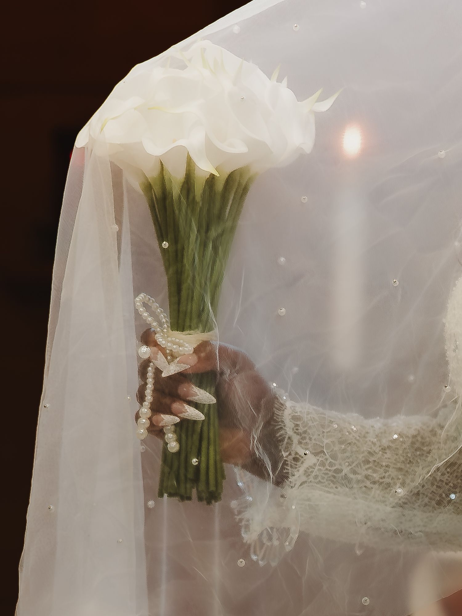 A bride holds a white calla lily bouquet wrapped in pearls beneath a sparkled veil.