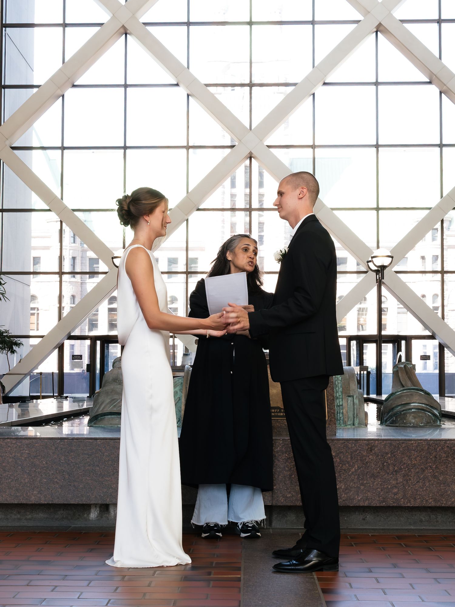 Couple exchanging vows at indoor wedding ceremony with officiant, bride in white gown, groom in black suit.