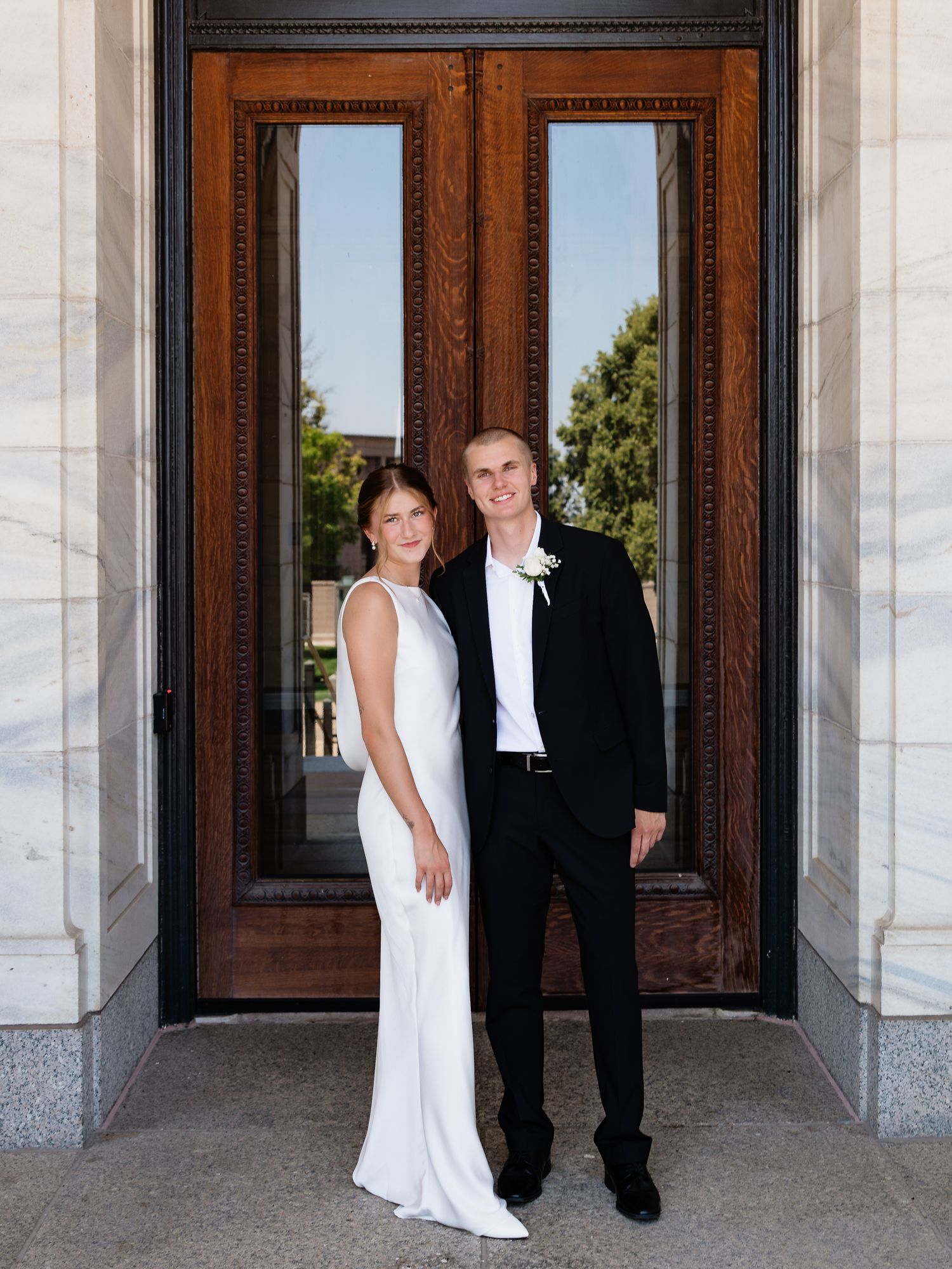 Bride in white gown and groom in black suit pose before ornate wooden doors at wedding venue.