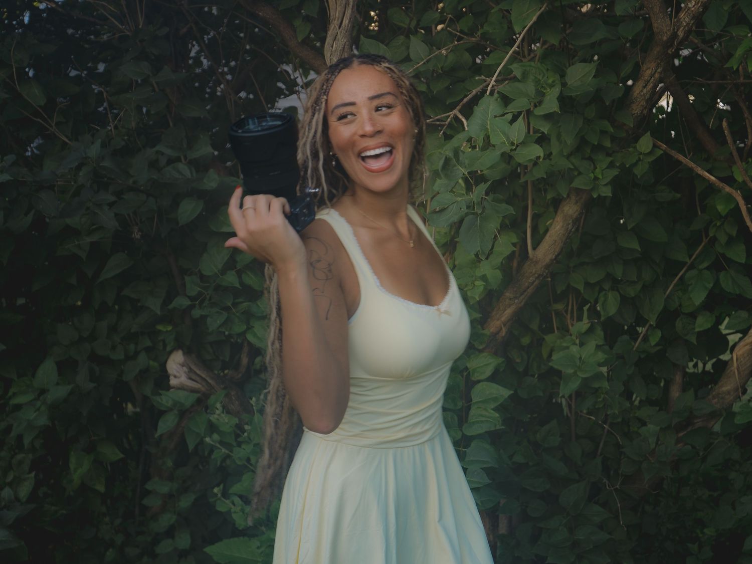 Joyful woman in white dress laughing while holding a camera among green leafy trees.