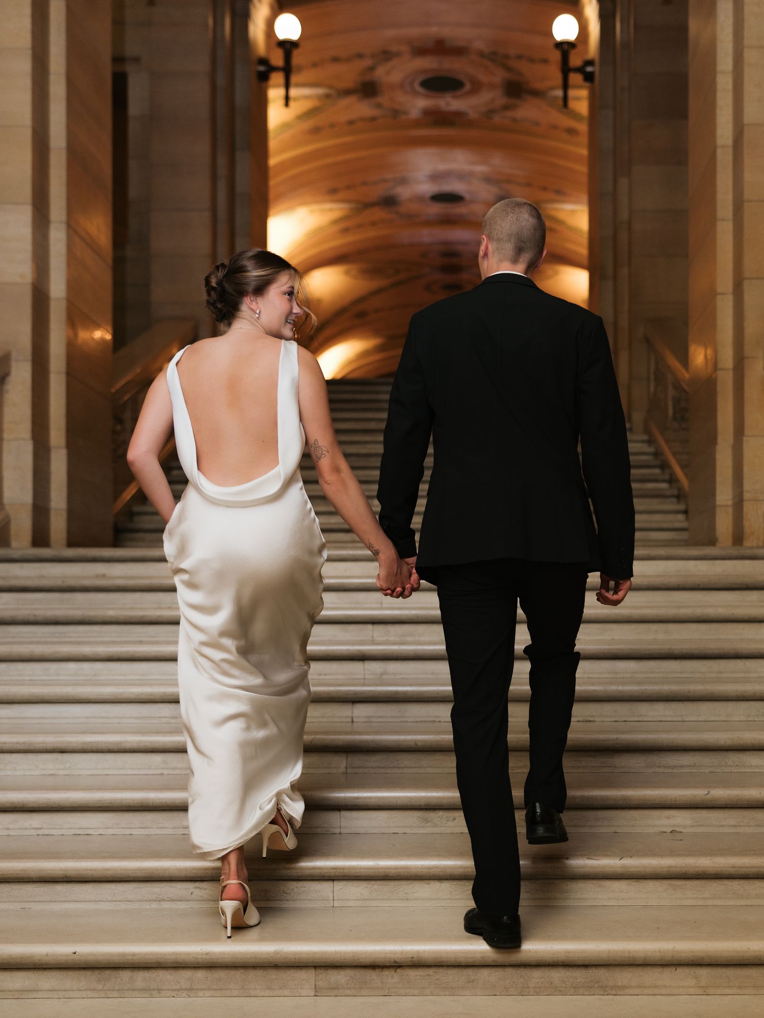 Bride in white satin backless gown and groom in black suit walk up grand marble staircase holding hands.