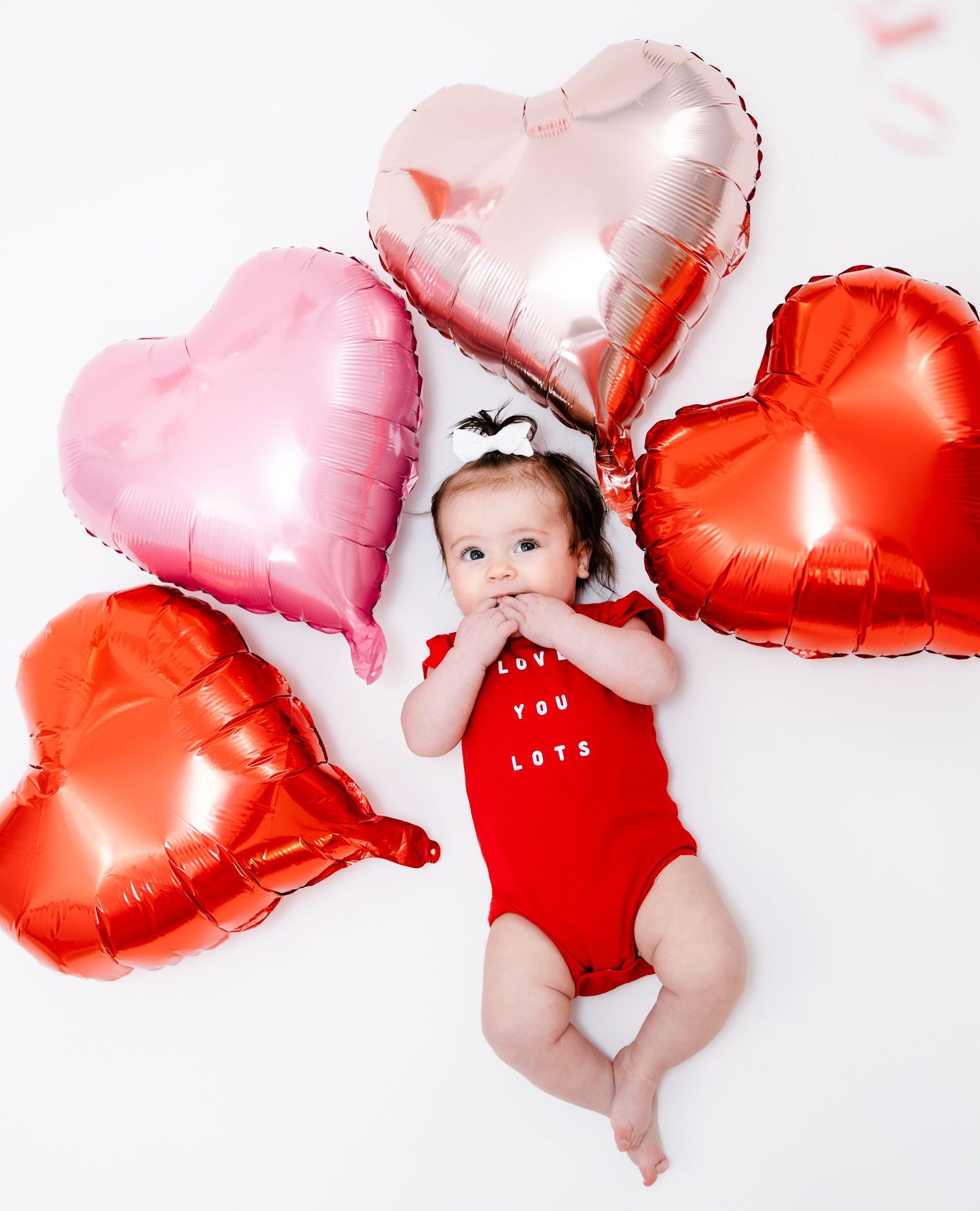 Sweet baby in red Love You Lots onesie lying among pink, rose gold, and red heart-shaped foil balloons.