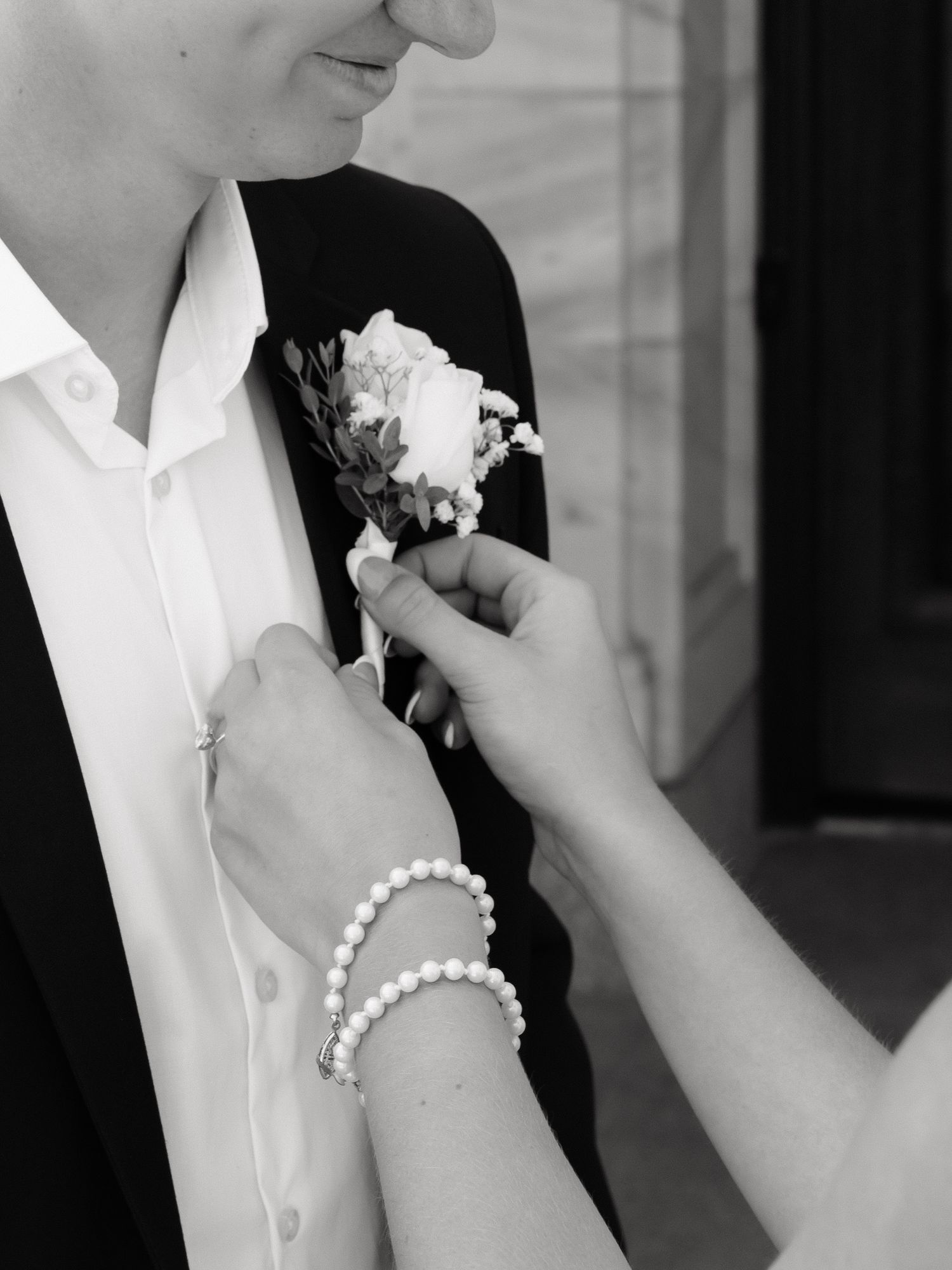 Hands with pearl bracelet pinning white floral boutonniere on groom's black suit lapel.