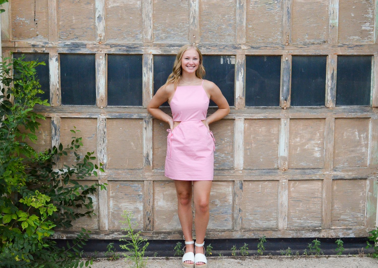 Smiling blonde woman in pink dress posing in front of rustic wooden garage doors.