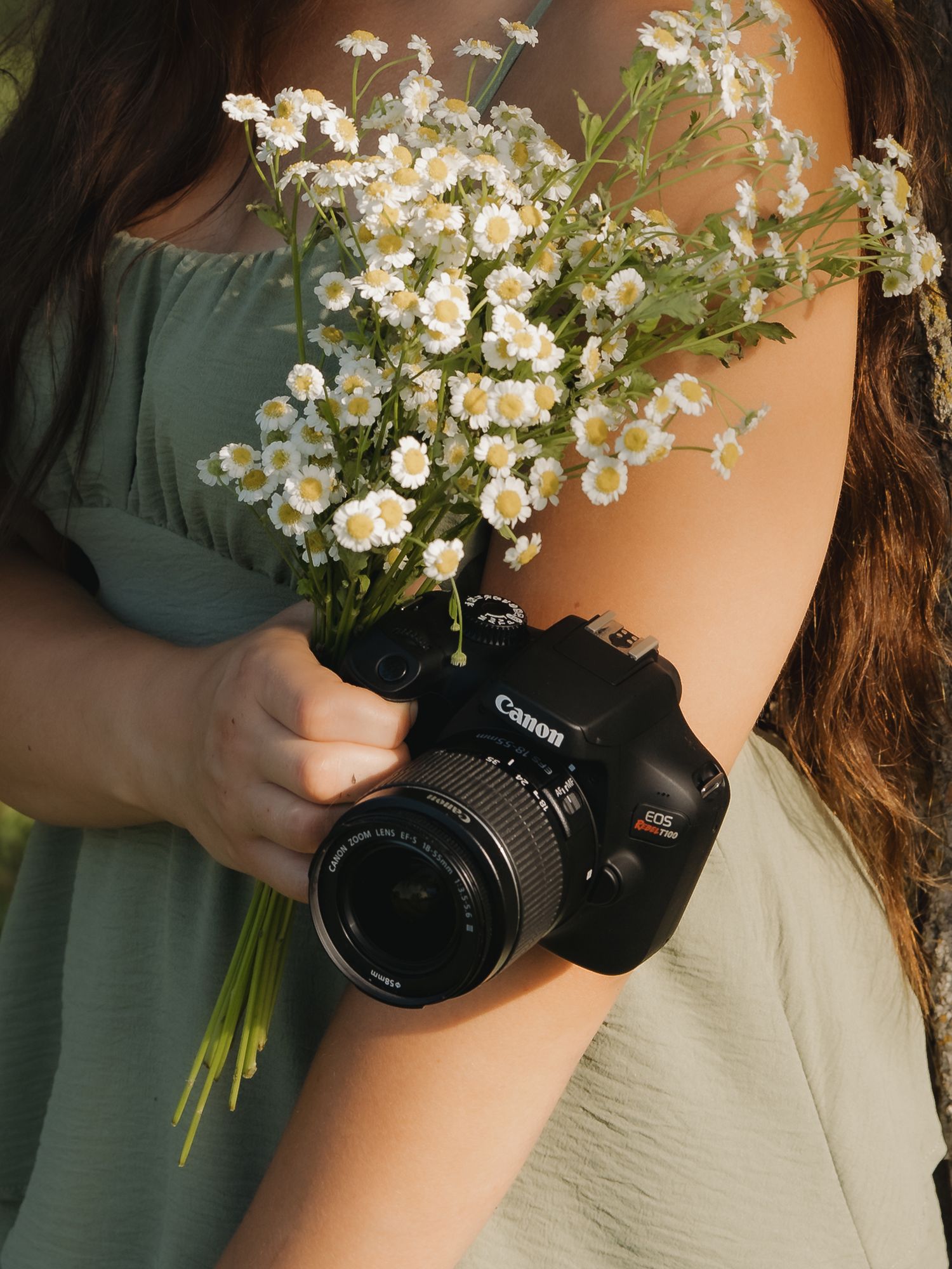 Woman in sage green dress holding Canon DSLR camera and white wildflower bouquet.
