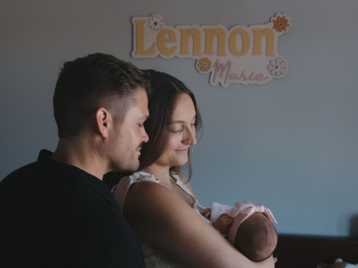 New parents tenderly holding their newborn baby under a wooden name sign reading Lennon.