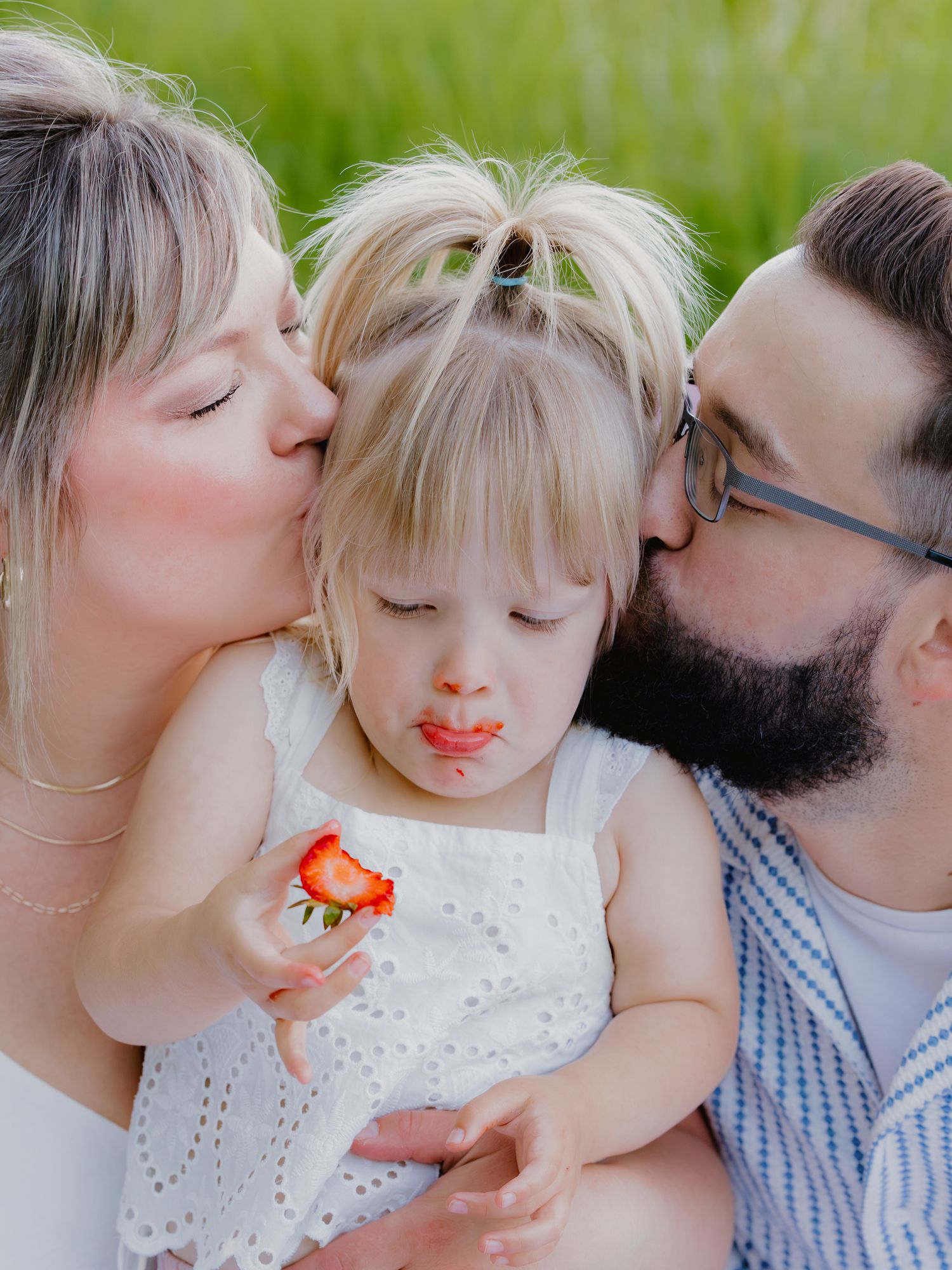 Parents kissing their young blonde toddler on the cheeks while she eats a strawberry.