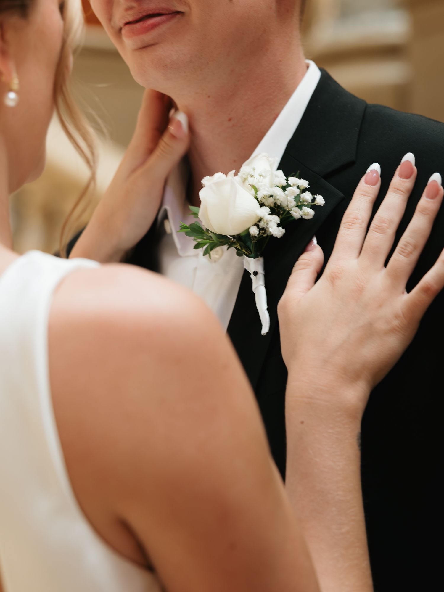 Bride placing hands on groom's chest near white rose boutonniere on black tuxedo at wedding.
