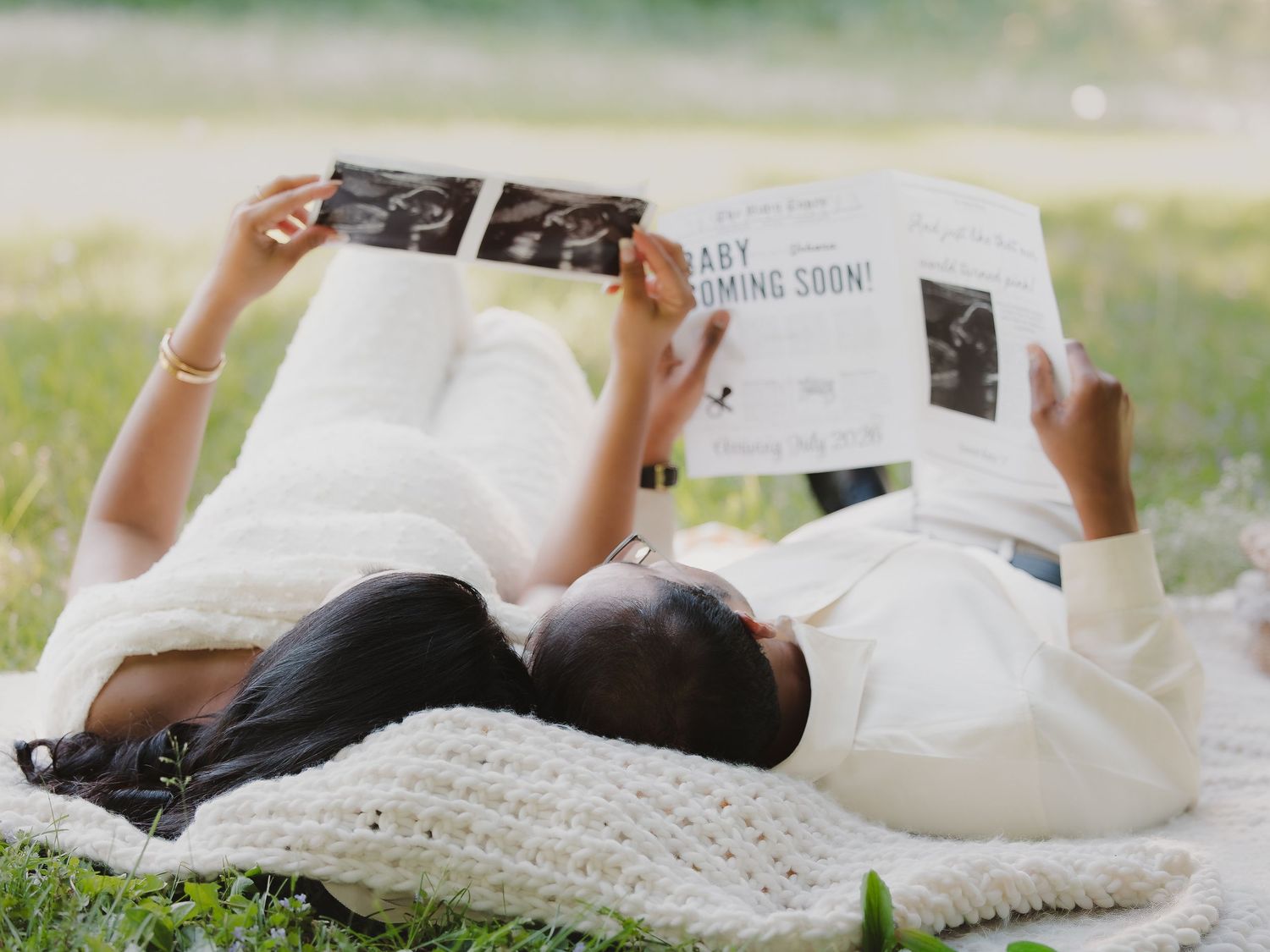 A couple lies on a blanket in a park, holding ultrasound photos and a baby announcement newspaper.