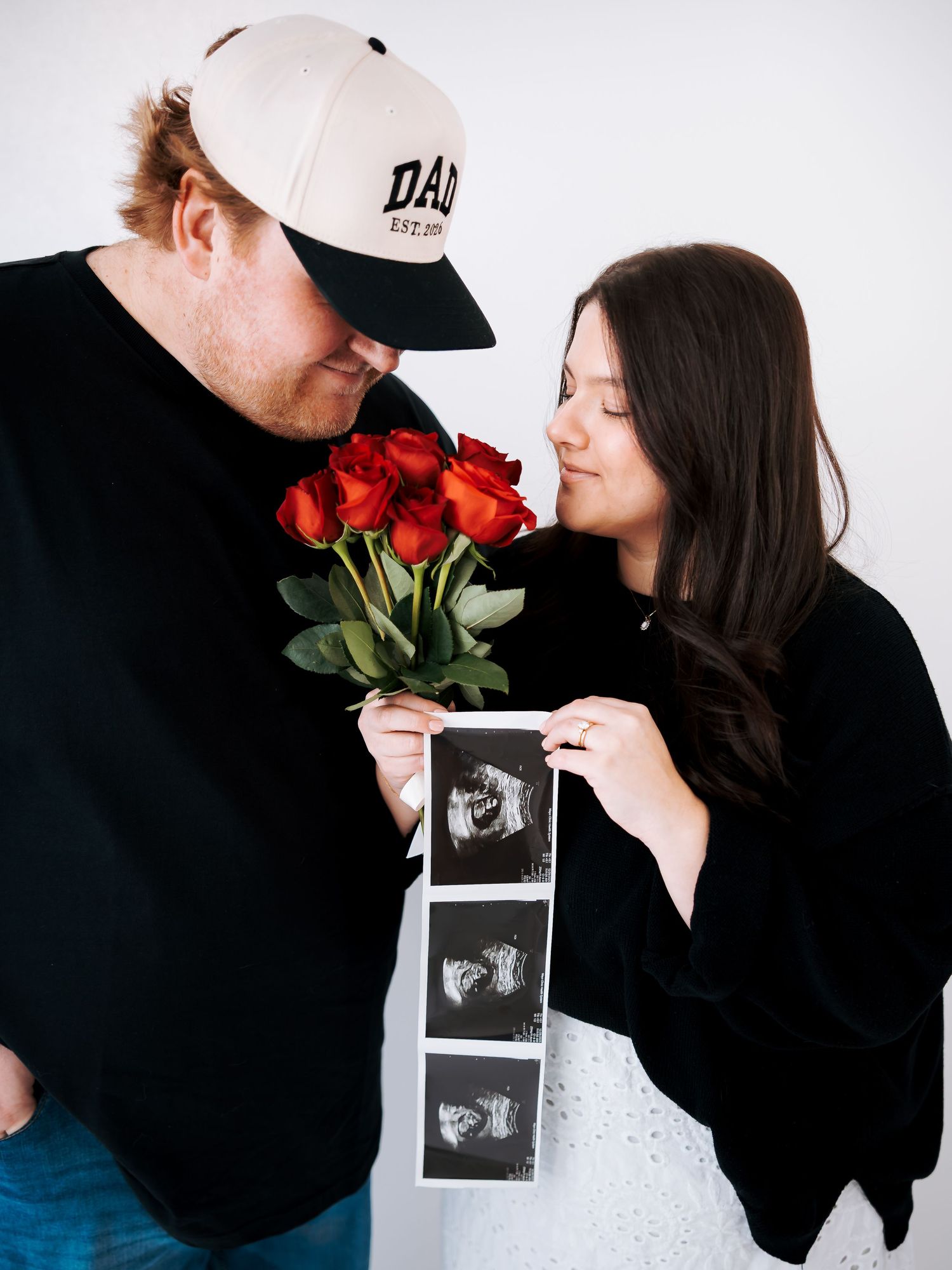 Expecting couple smelling red roses while holding ultrasound photos in pregnancy announcement photo.