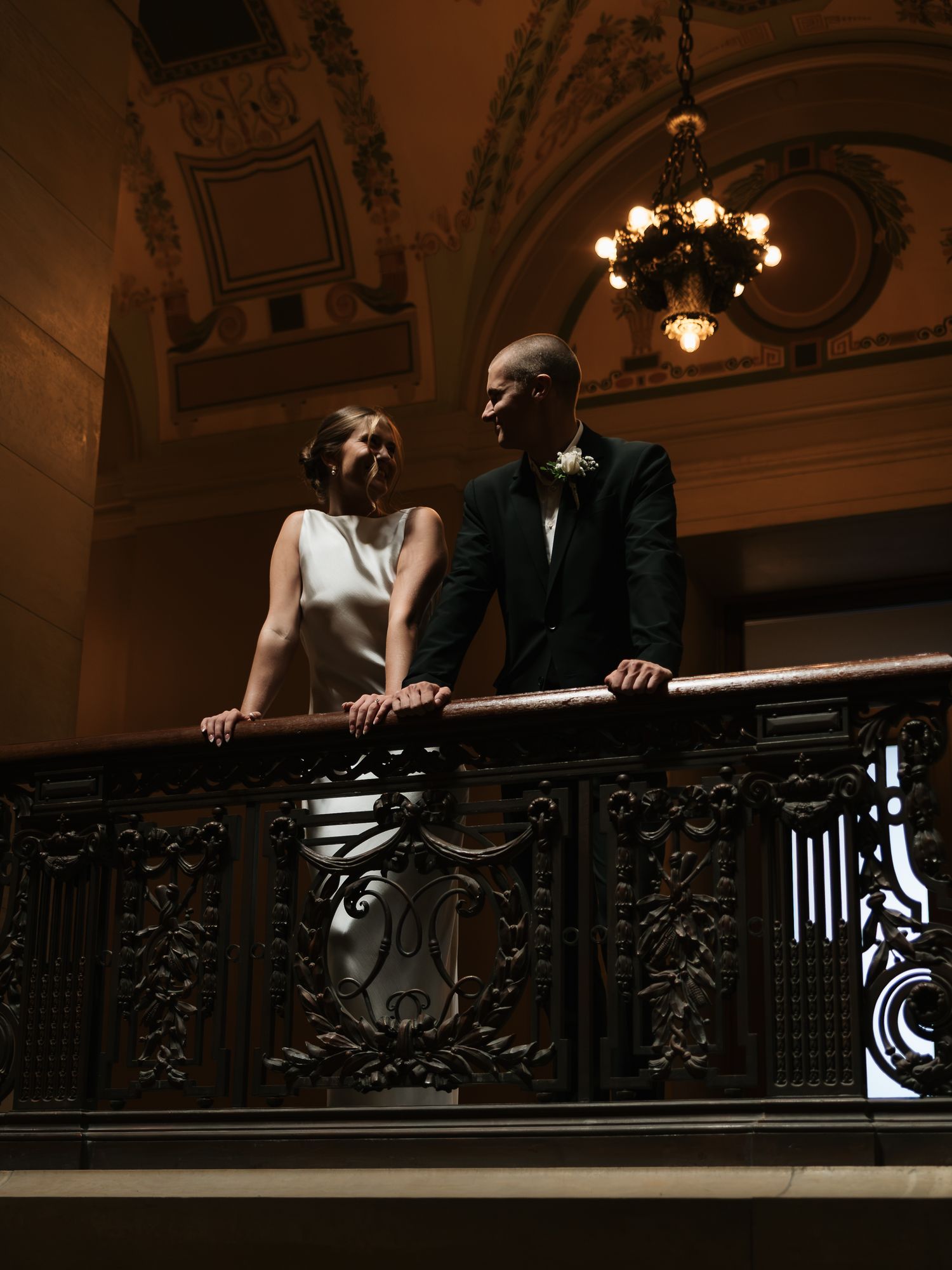 Newlyweds standing on ornate carved balcony railing in grand historic building.