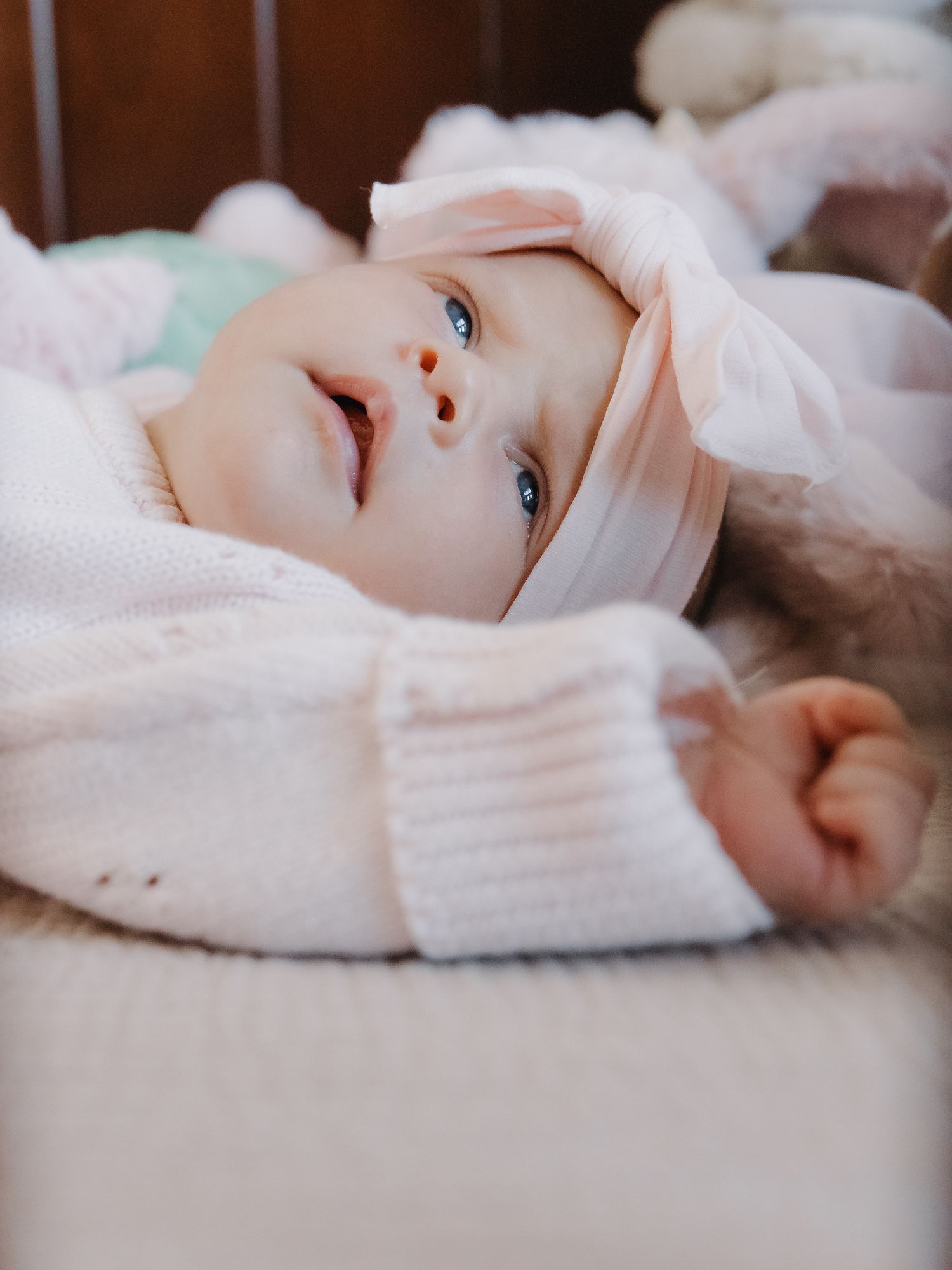 Blue-eyed baby in pink knit sweater and bow headband lying on soft blanket looking up sweetly.