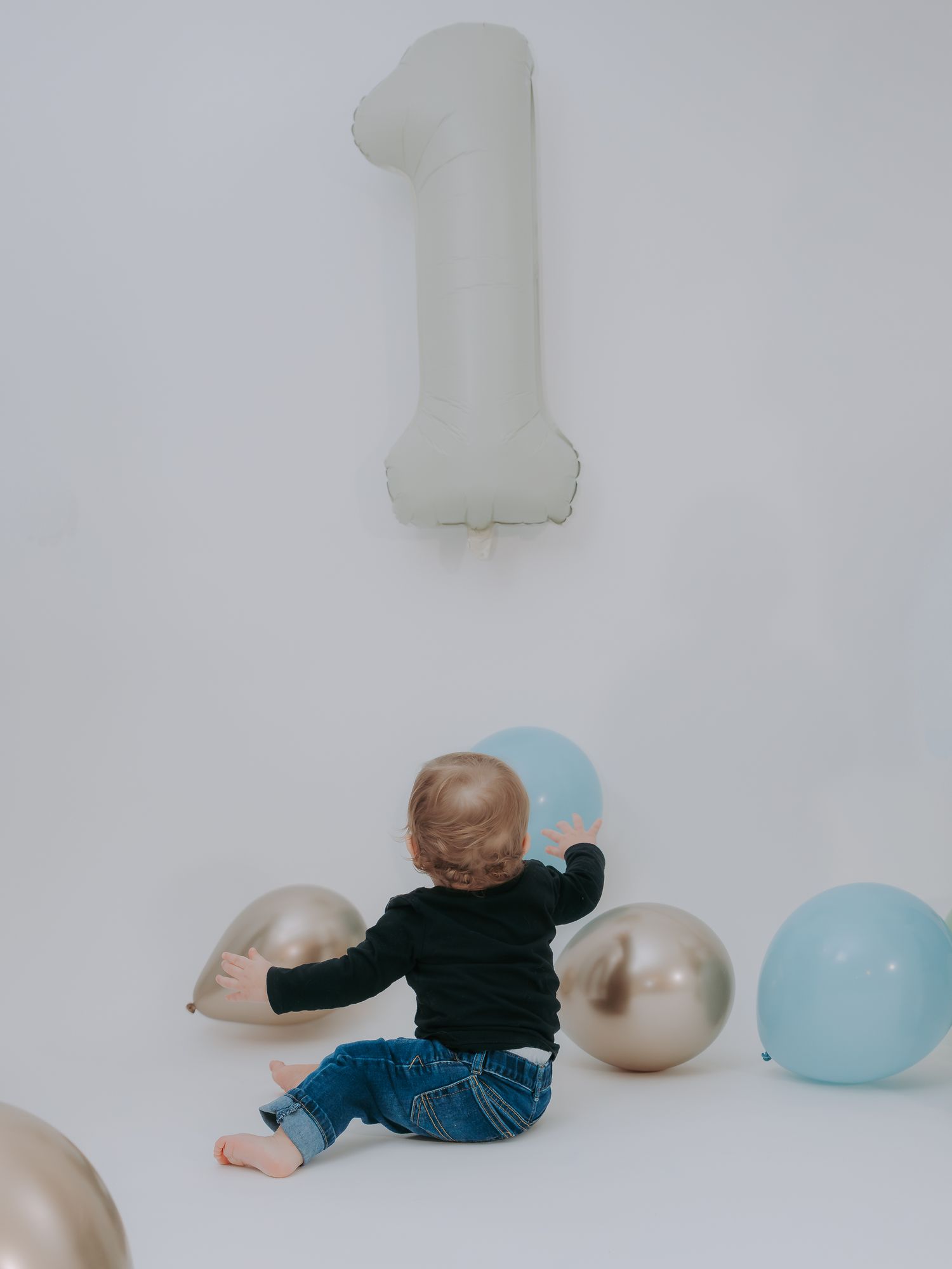 Baby in black shirt and jeans reaching for blue balloon near silver balloons and white number one foil balloon.