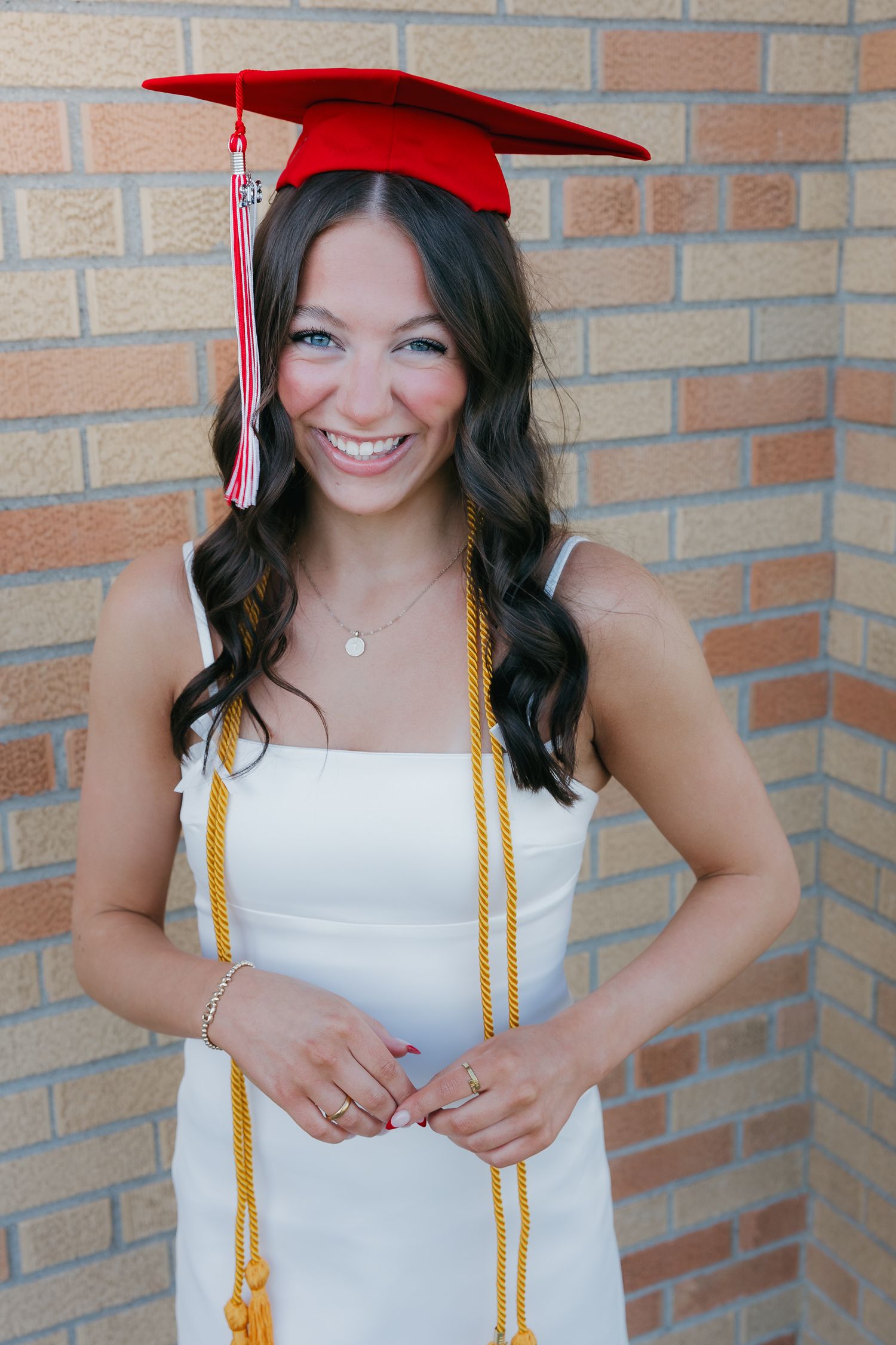 Smiling brunette woman in white dress wearing red graduation cap and gold honor cords.