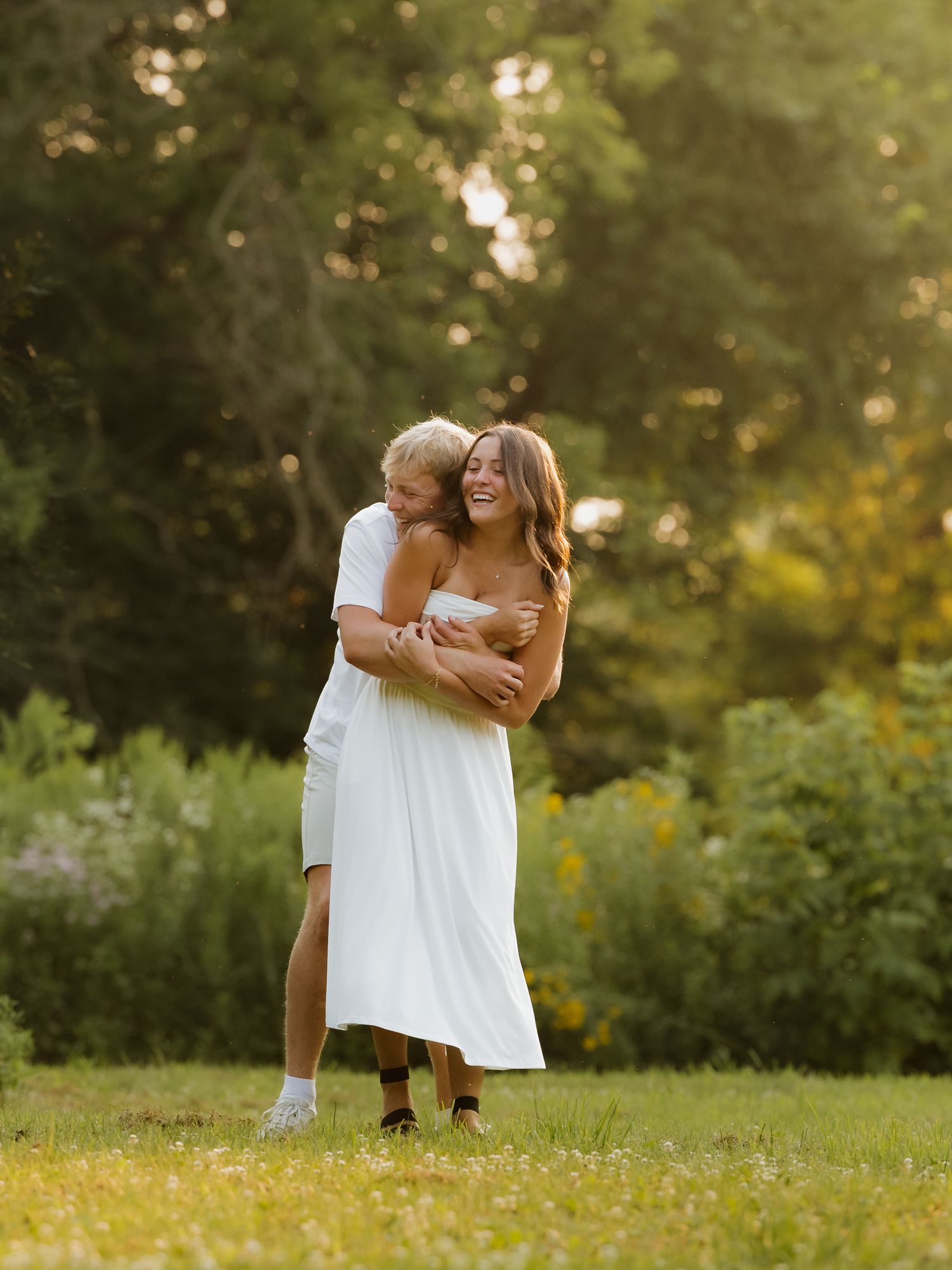 Couple embracing and laughing in a sunlit green meadow, woman in white dress, golden hour outdoor portrait.