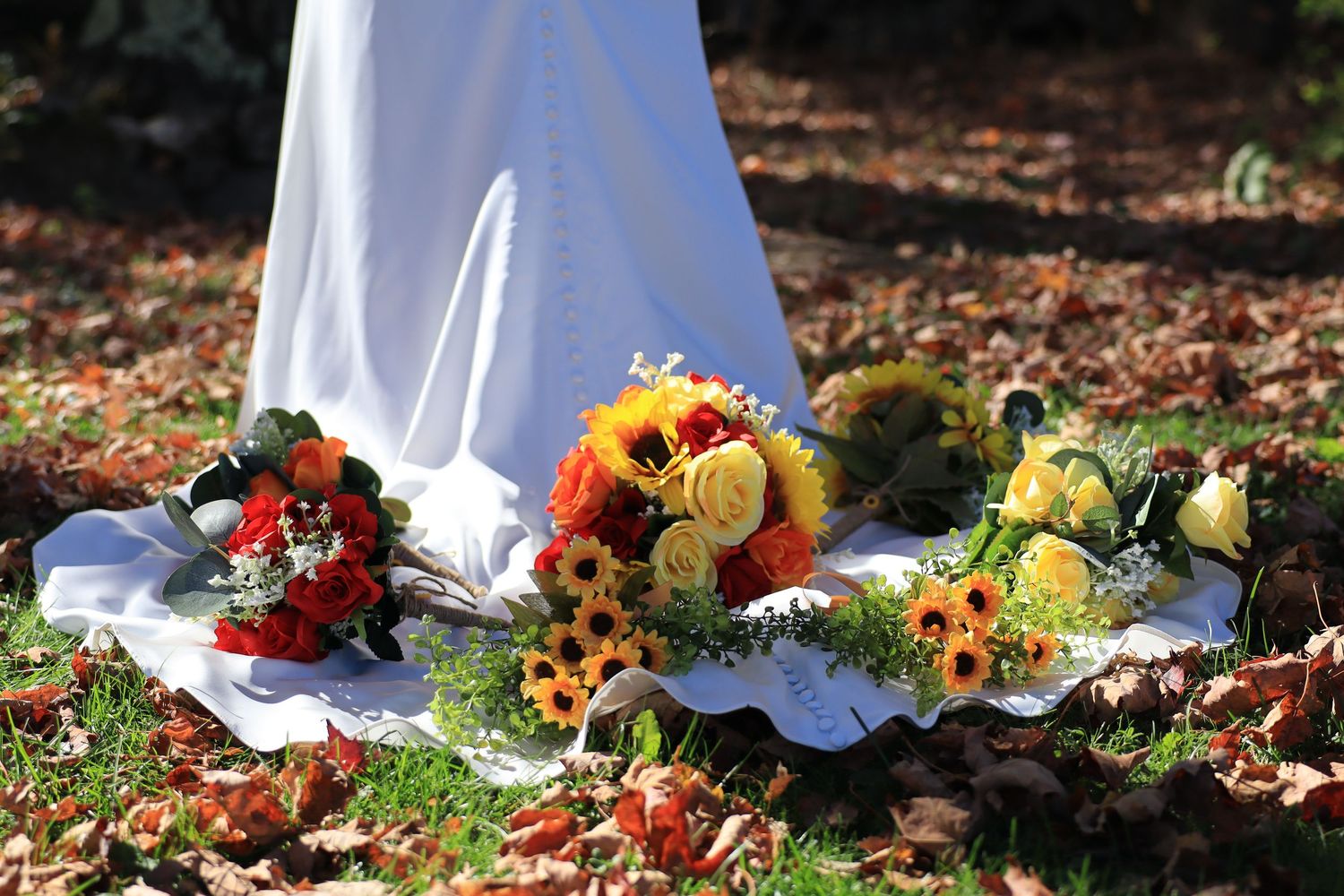 Autumn wedding bouquets of sunflowers and red roses arranged on white fabric among fallen leaves.