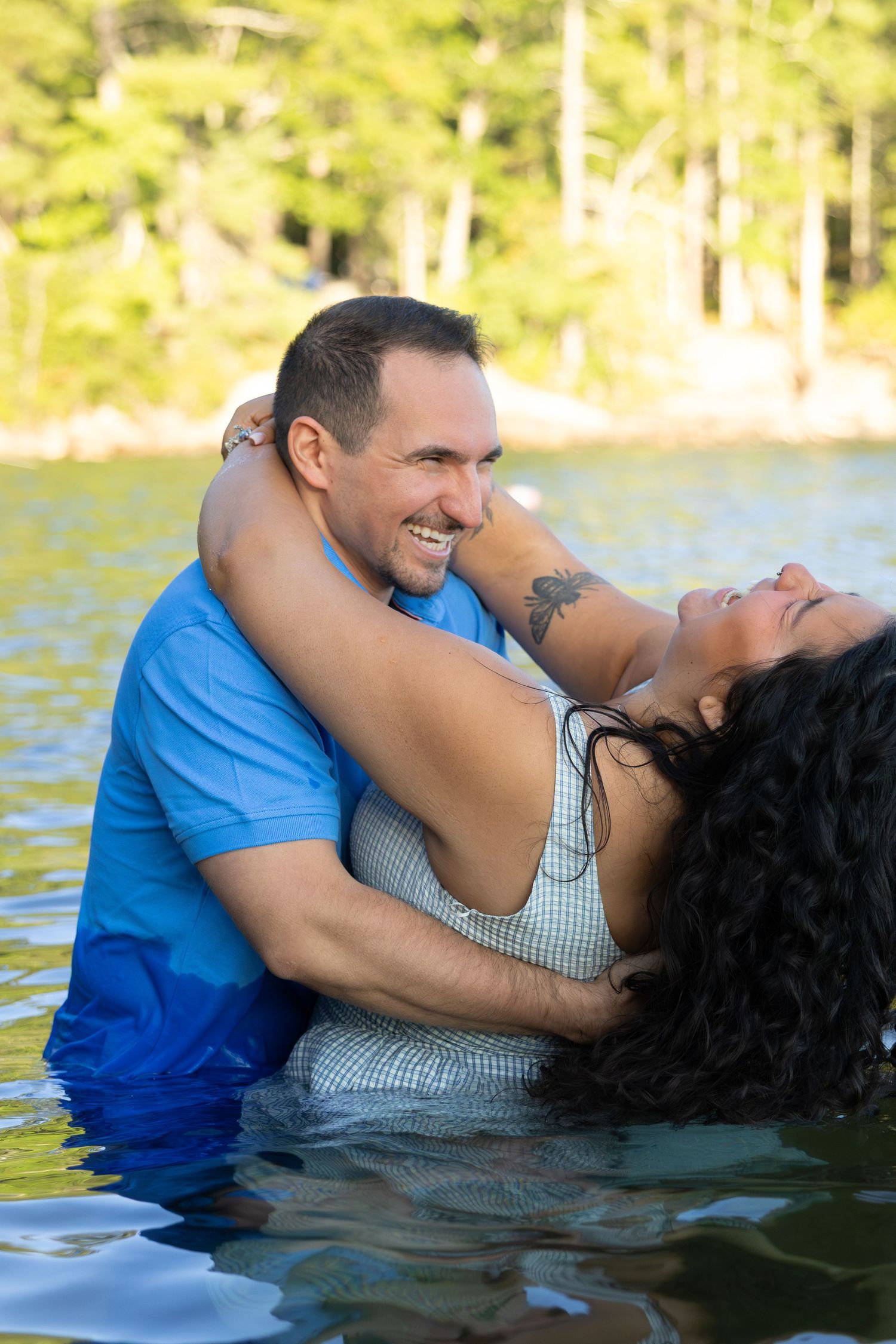Couple laughing and embracing in a lake, woman with tattoo leaning back joyfully, trees reflected in water behind them.