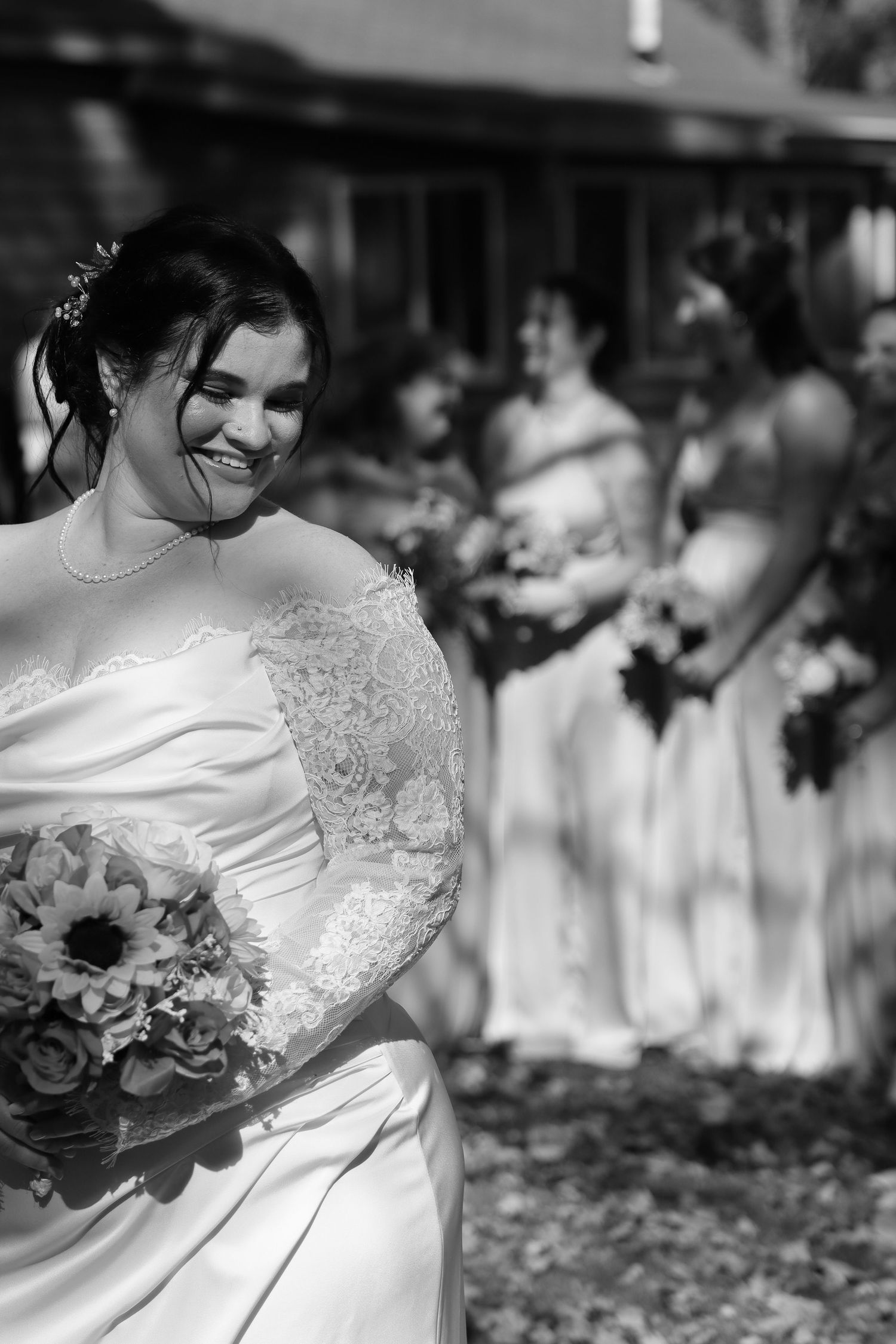 Smiling bride in lace wedding gown holds sunflower bouquet with bridesmaids in background, black and white.