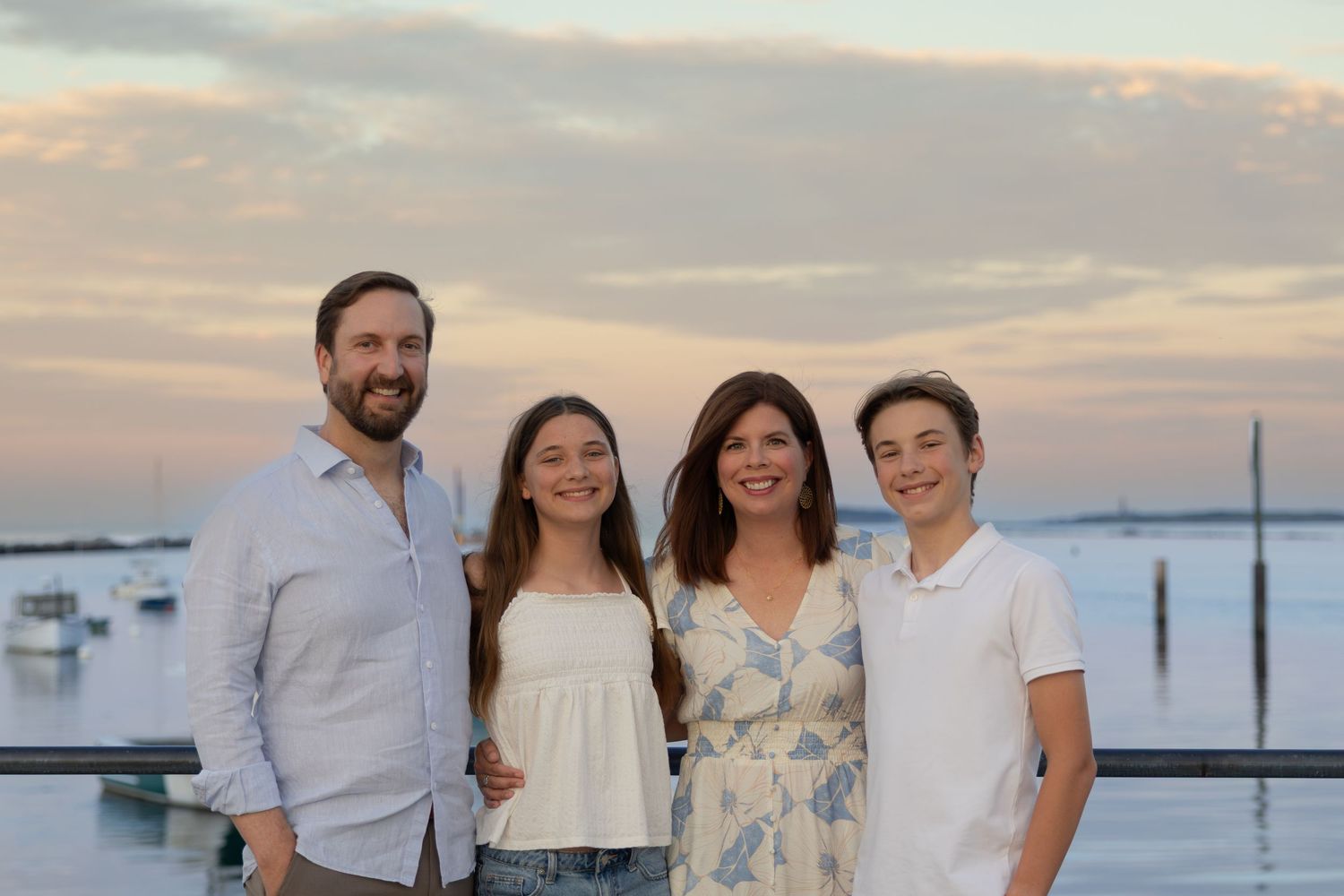 Family of four smiling together at waterfront during sunset with boats in background.