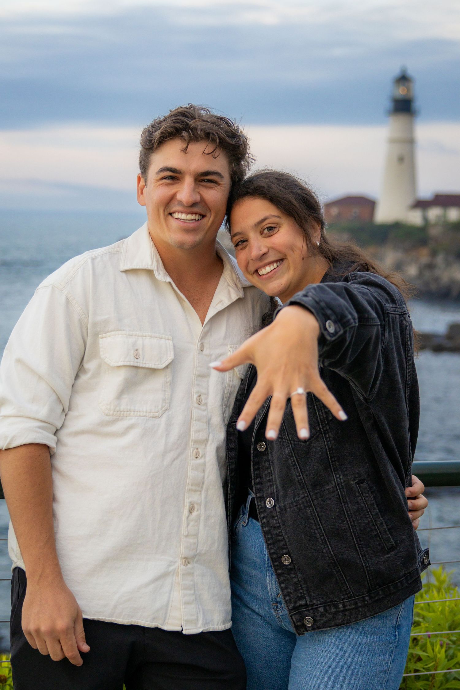 Happy engaged couple smiling near lighthouse at dusk, woman showing off engagement ring to camera.