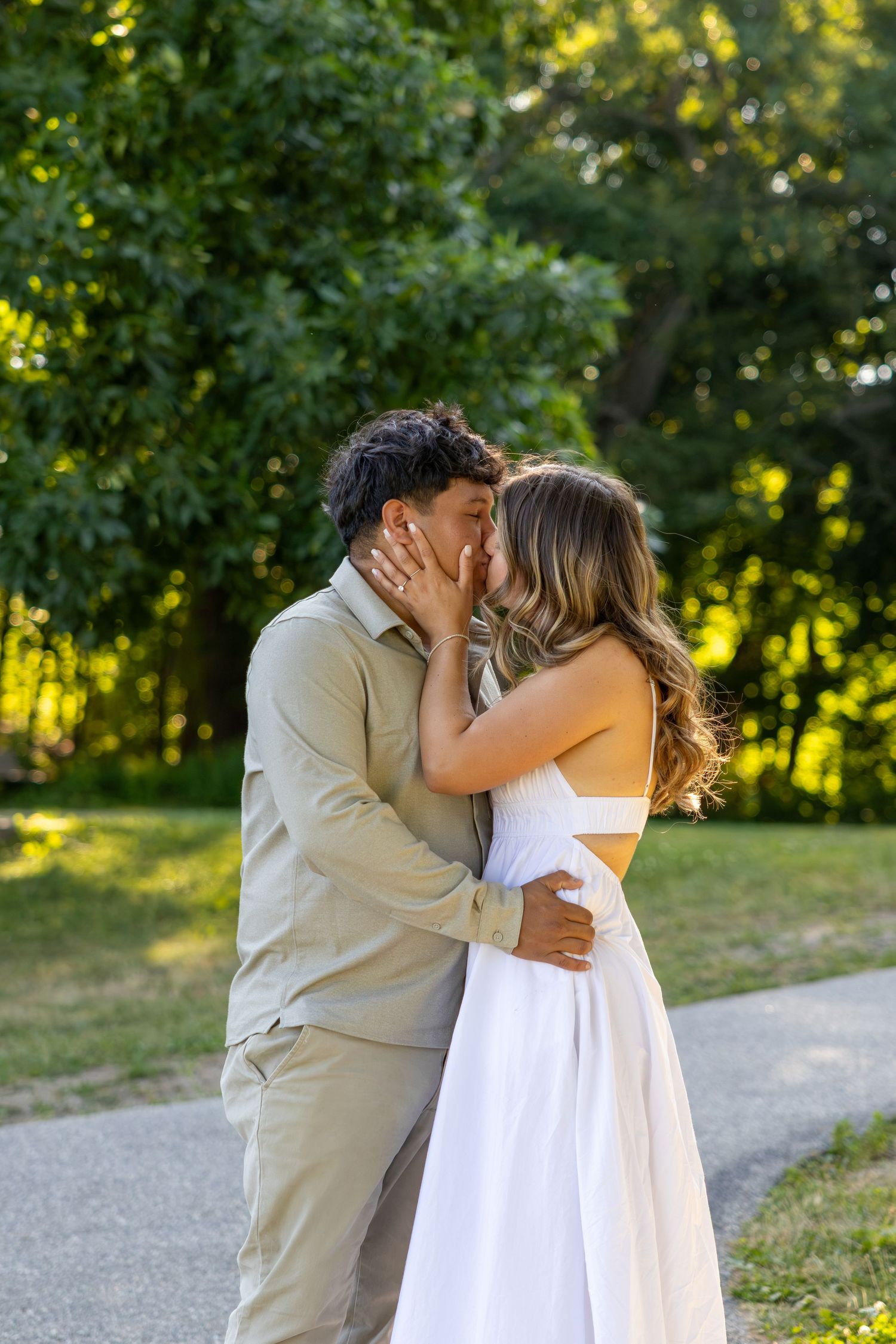 Couple sharing a romantic kiss outdoors in a lush green park, woman in white dress, man in beige outfit.