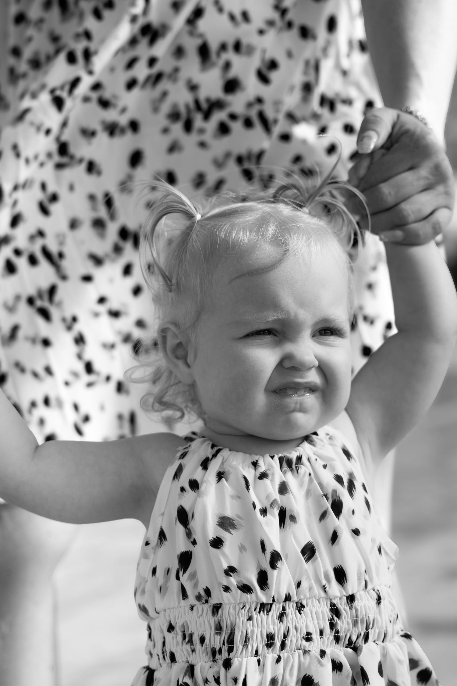 Black and white photo of a toddler girl in a spotted dress making a funny face while an adult holds her hand.