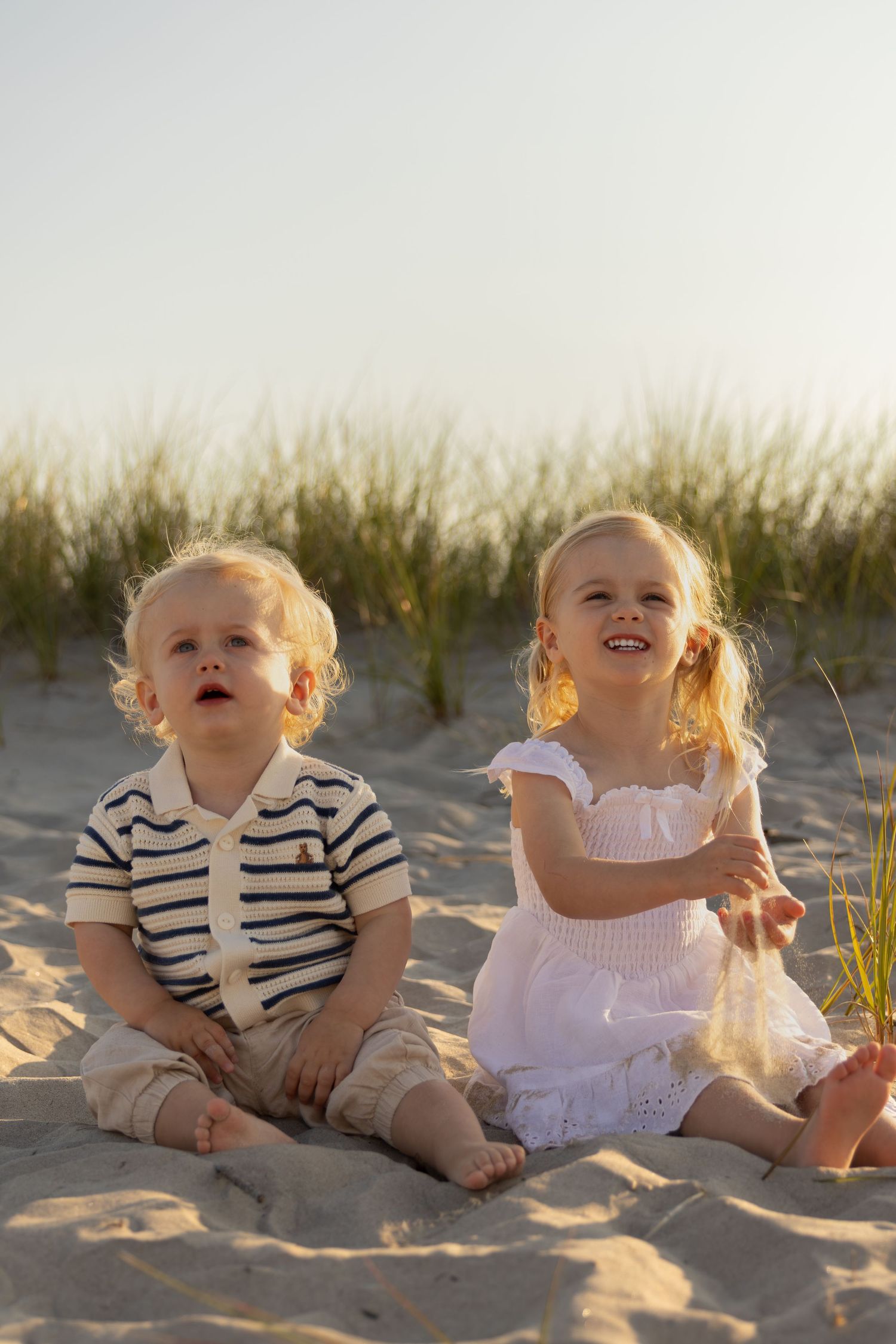 Two young blonde toddlers sitting barefoot on sandy beach, boy in striped shirt and girl in white dress, smiling outdoors.