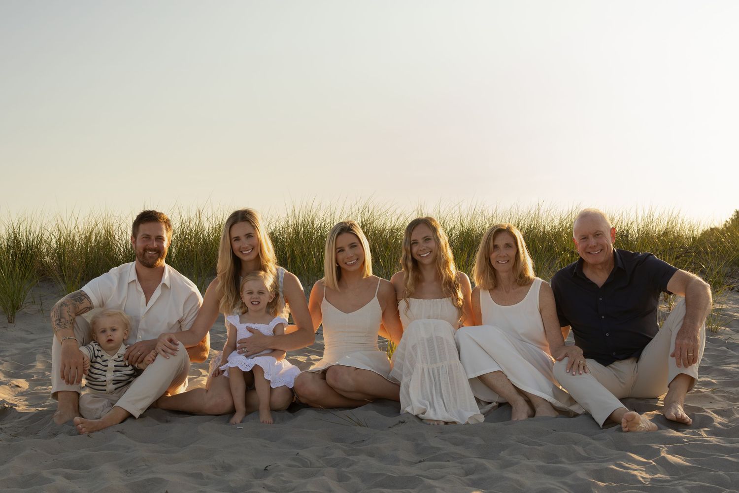 Family group photo on sandy beach at sunset, adults and toddlers dressed in white and navy outfits.