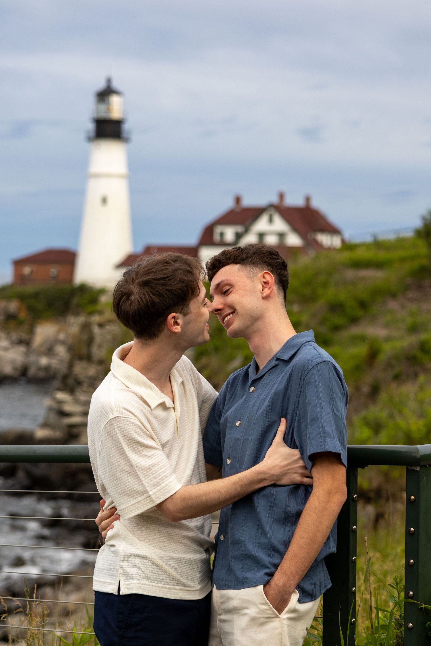 Two men embracing and smiling at each other with a white lighthouse and ocean in the background.