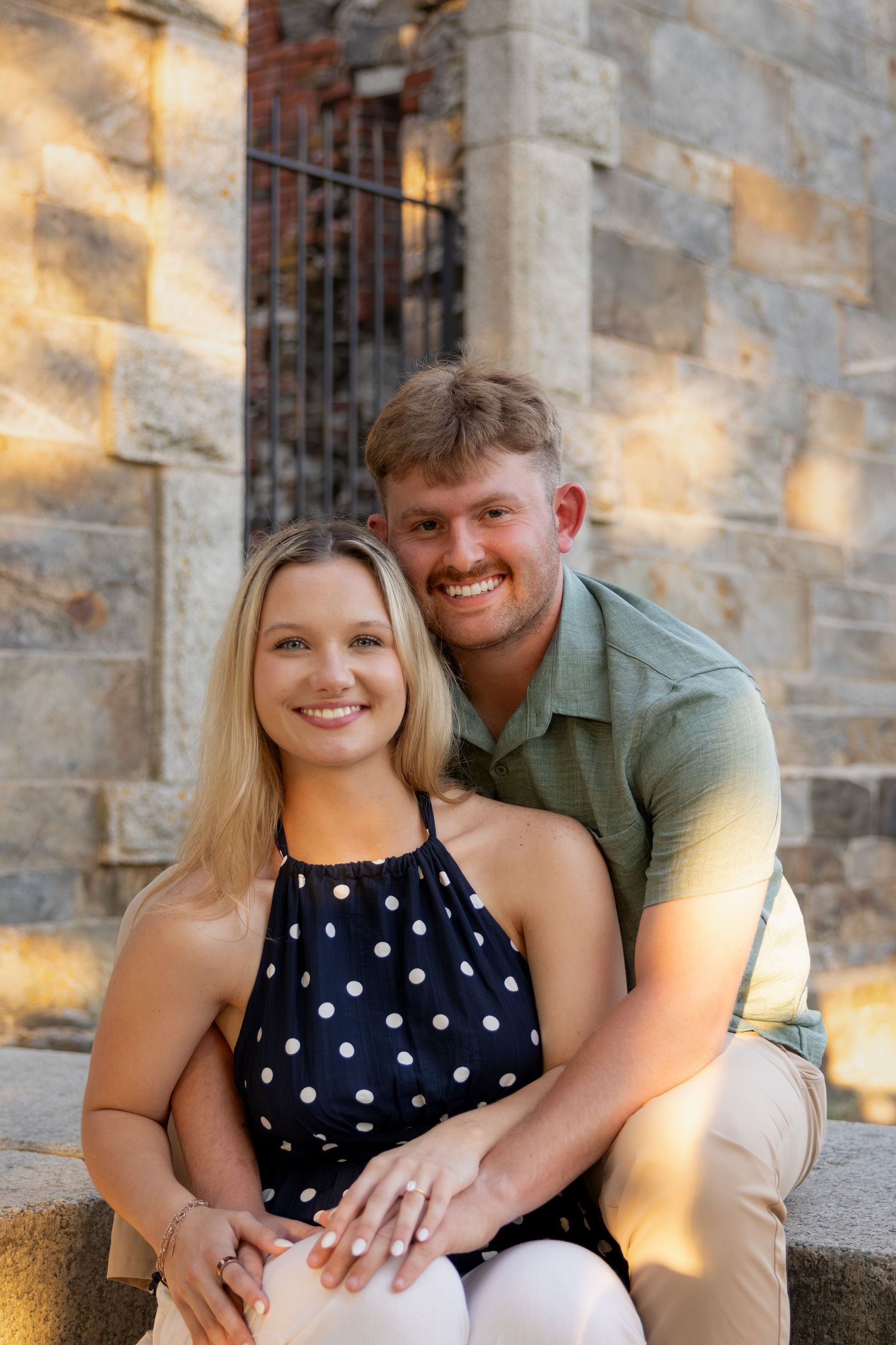Smiling couple sitting together outdoors against a stone wall, woman in polka dot top, man in green shirt.