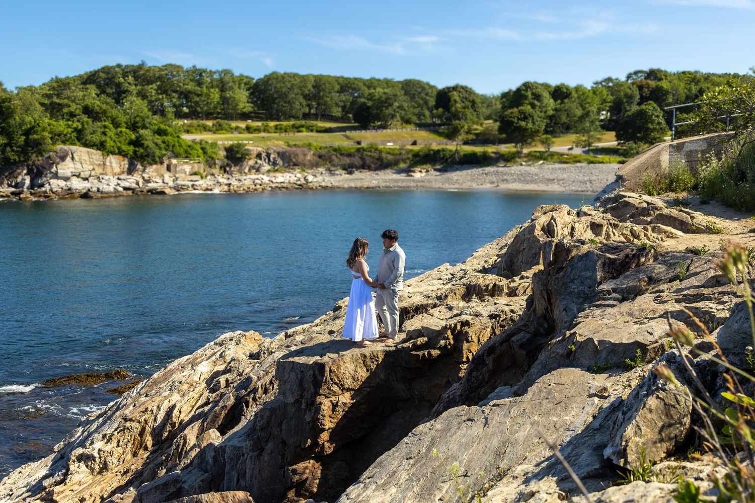 Couple in white attire standing on rocky coastal cliffs with calm blue water and lush green trees in background.