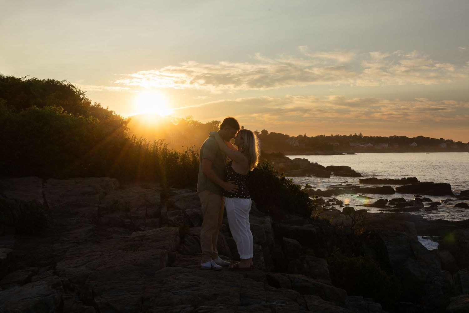Couple silhouetted against a golden sunset on rocky coastal shore, sharing a romantic embrace by the ocean.